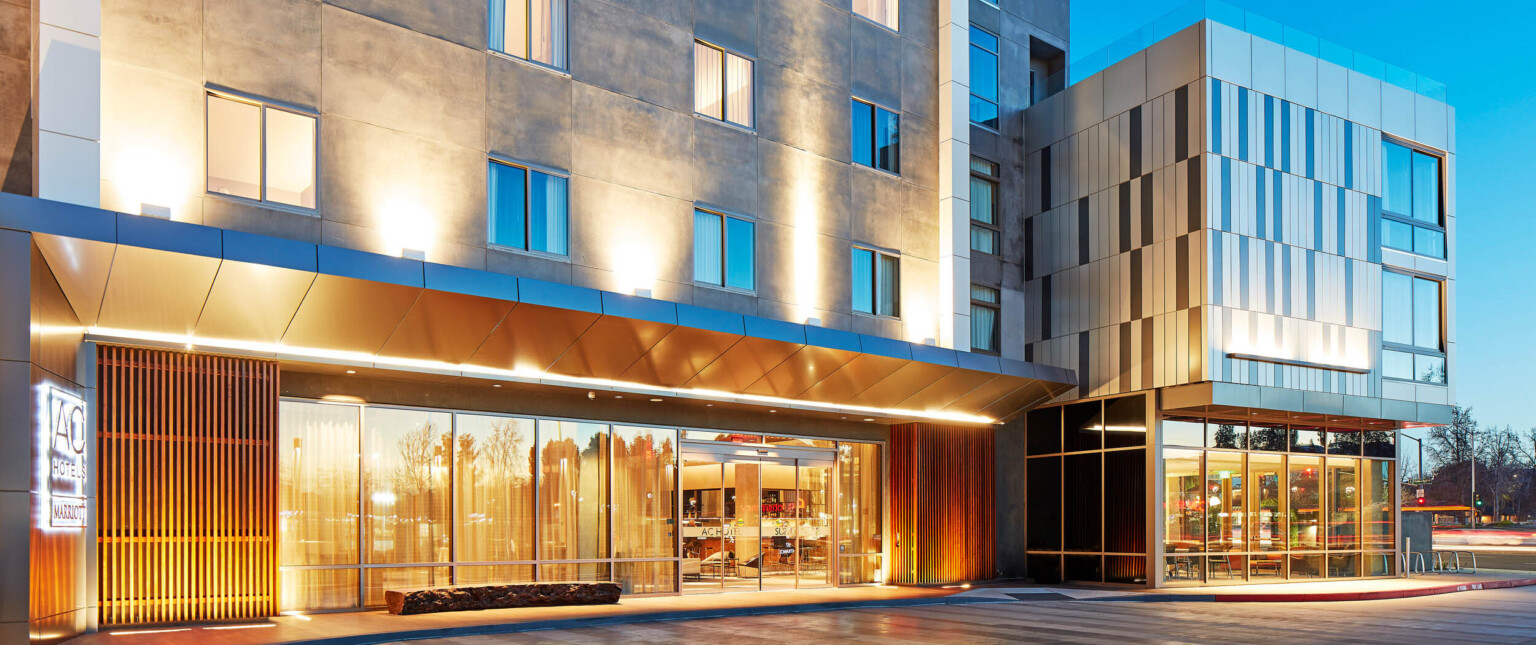 Sliding door entry to hotel illuminated from recessed light in overhang. Stone building with wood panels at bottom corners following Marriott brand architecture