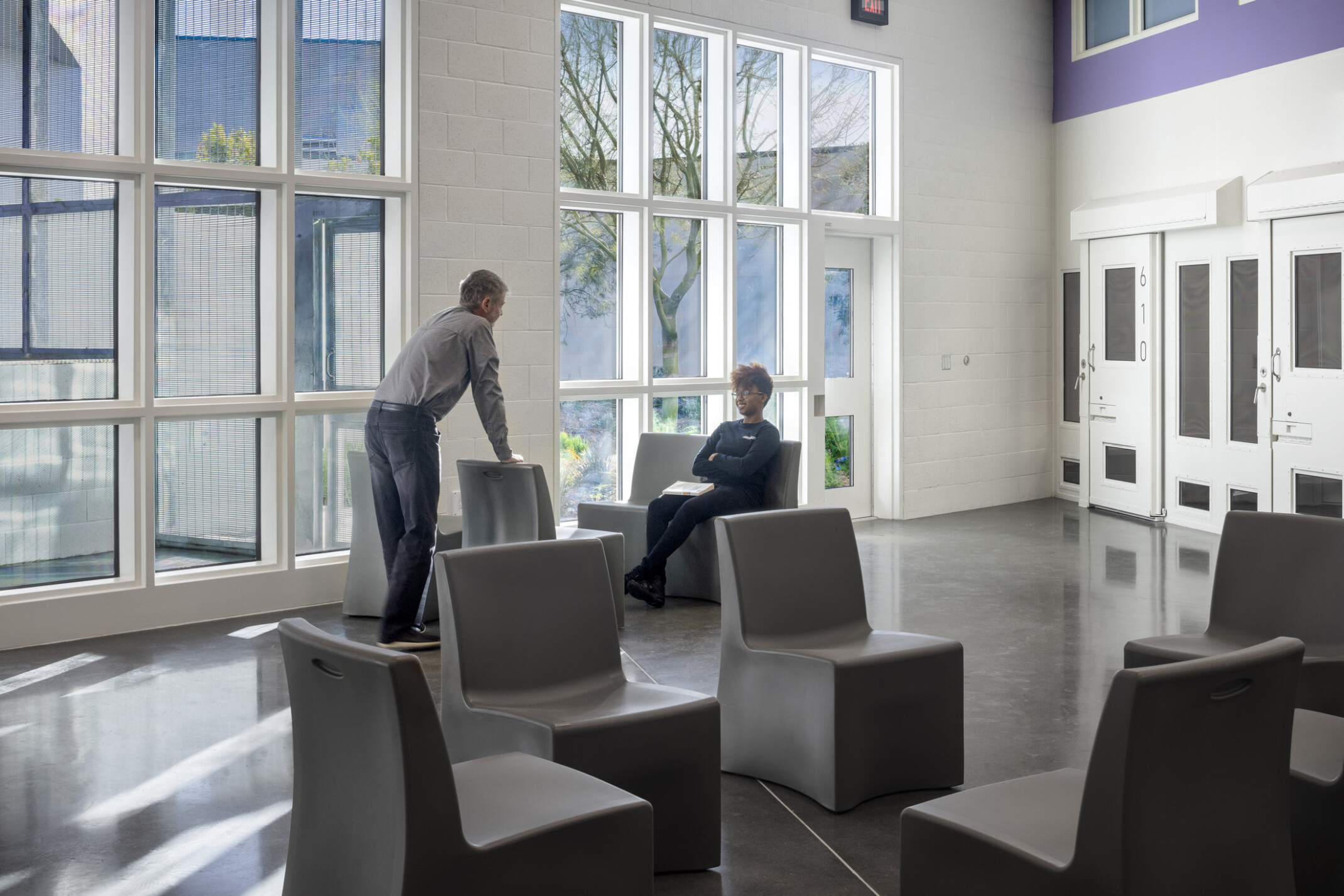 An interior photo of two people talking inside a communal area of the building, double-height windows, purple accent wall