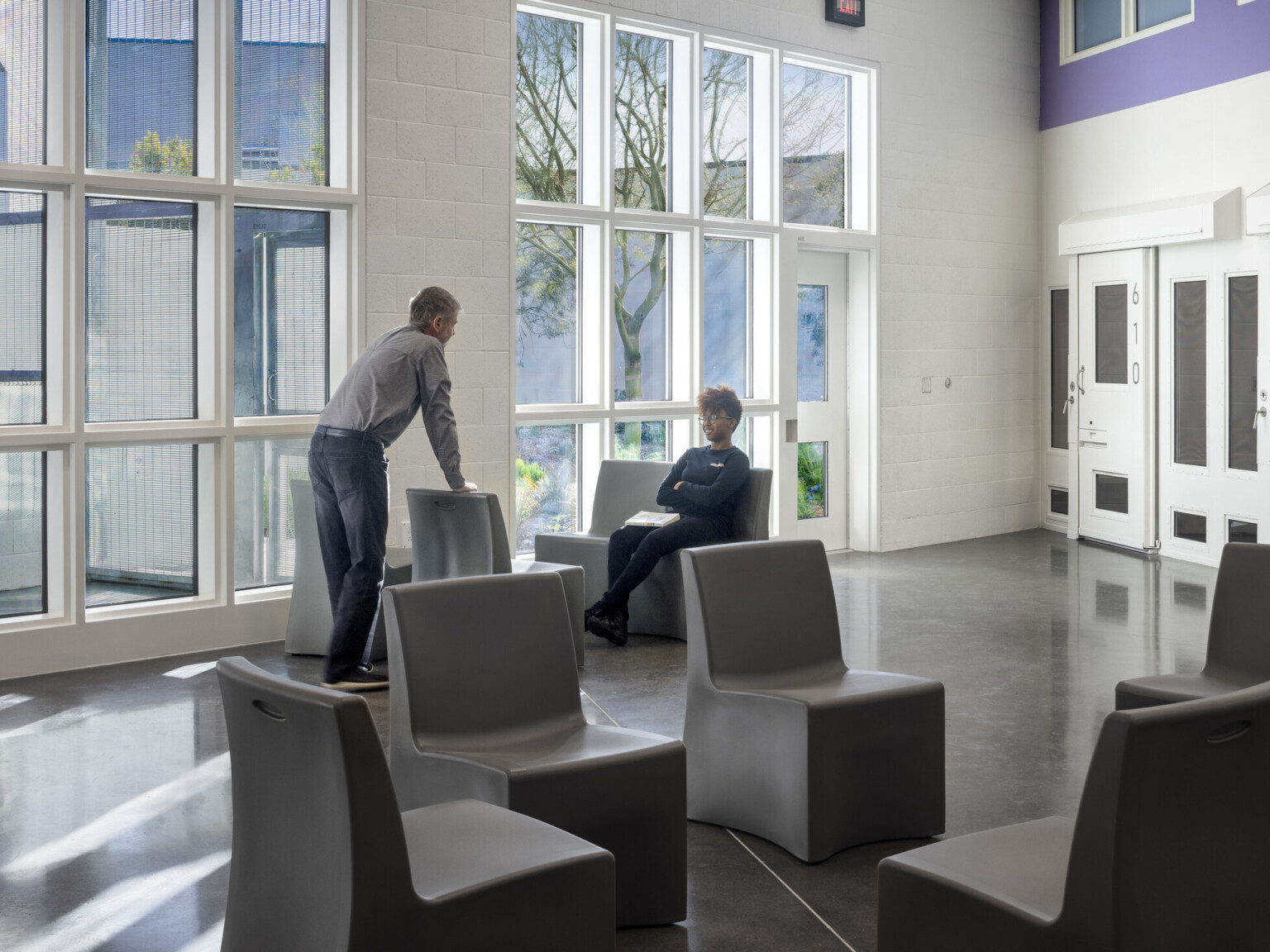 An interior photo of two people talking inside a communal area of the building, double-height windows, purple accent wall