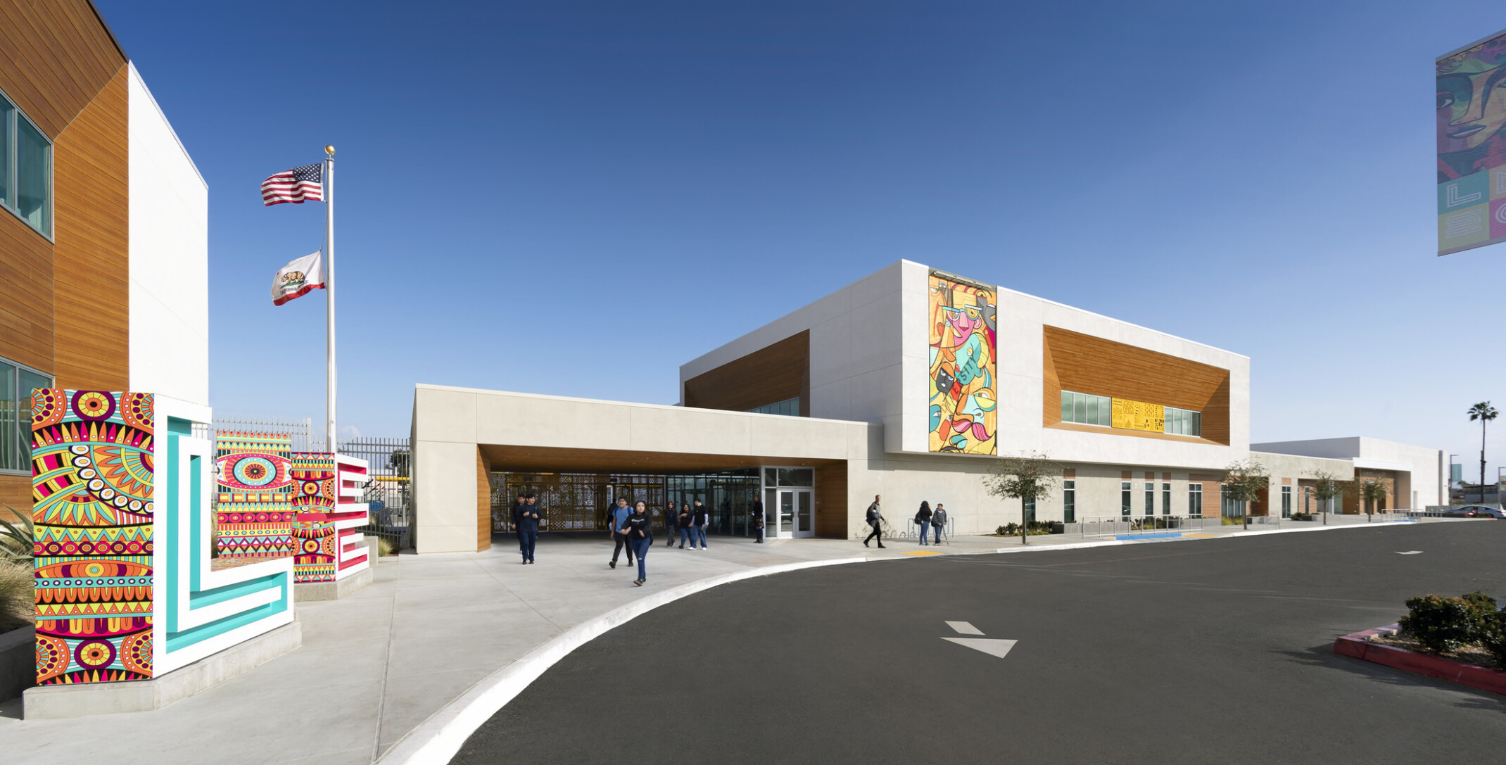 exterior view of building entrance, parking lot, brightly colored murals painted on two beige concrete and mass timber paneled façades, flagpole flying two flags