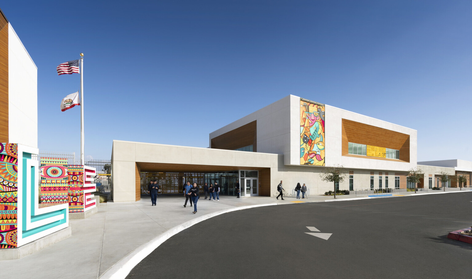 exterior view of building entrance, parking lot, brightly colored murals painted on two beige concrete and mass timber paneled façades, flagpole flying two flags