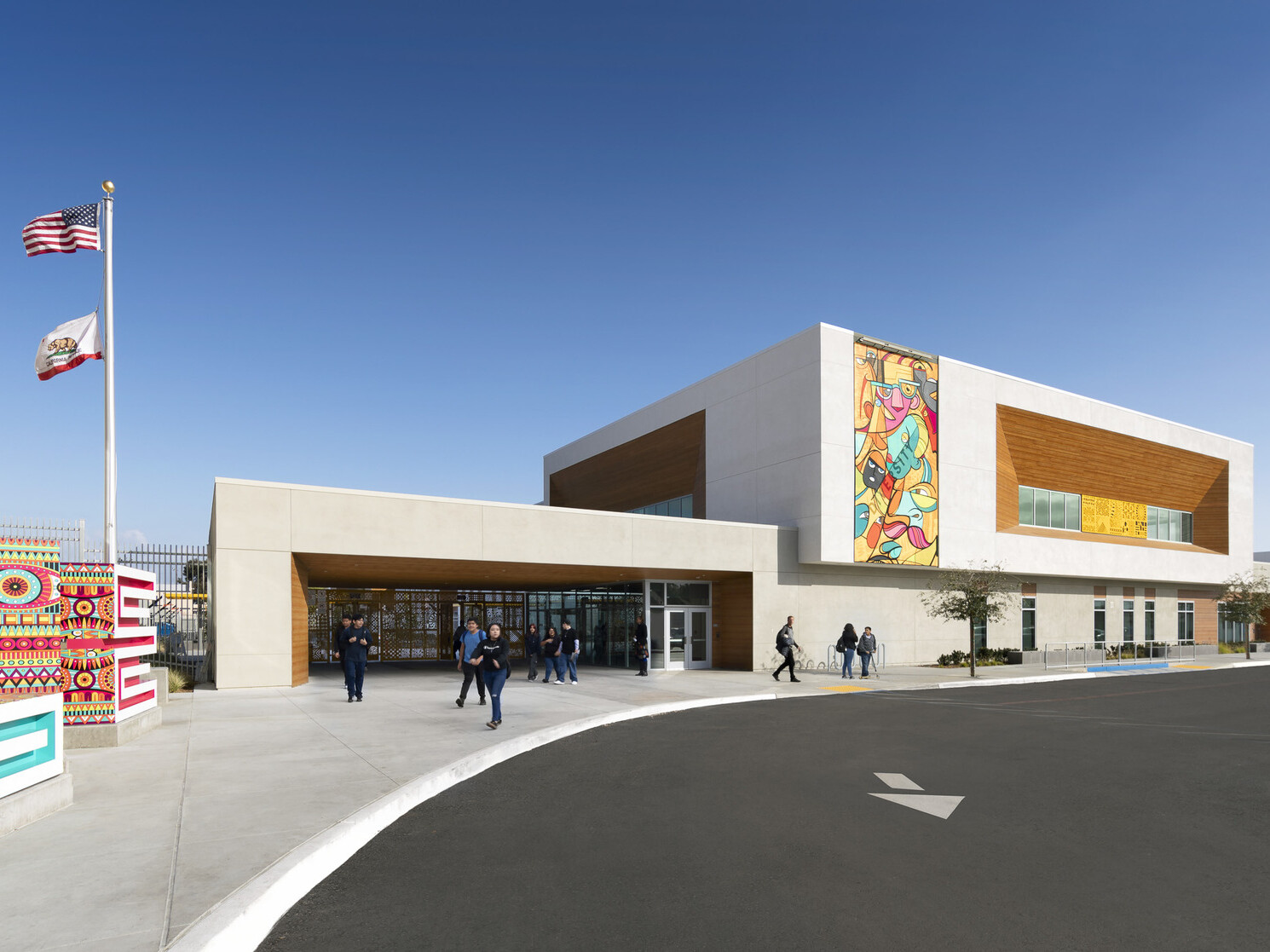 exterior view of building entrance, parking lot, brightly colored murals painted on two beige concrete and mass timber paneled façades, flagpole flying two flags exterior view of building entrance, parking lot, brightly colored murals painted on two beige concrete and mass timber paneled façades, flagpole flying two flags