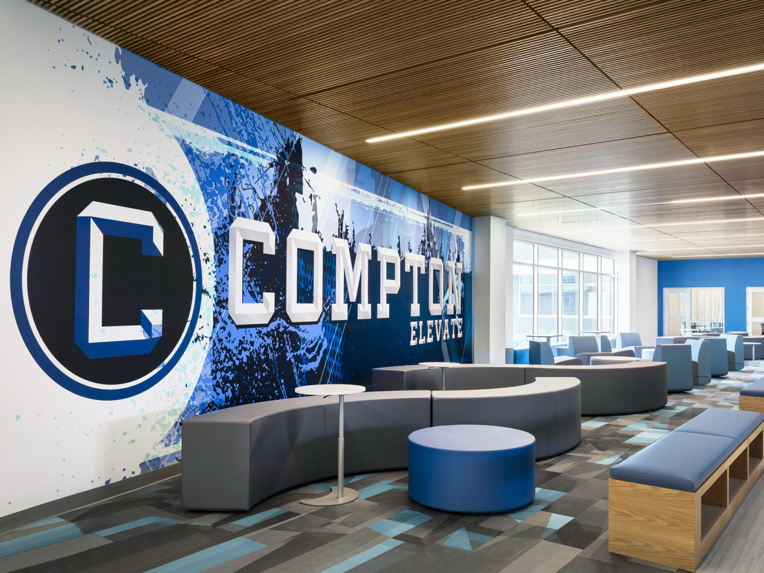 Common area of Compton High School; blue and white graphic design feature wall with "Compton" highlighted; community based-design seating Common area of Compton High School; blue and white graphic design feature wall with "Compton" highlighted; community based-design seating