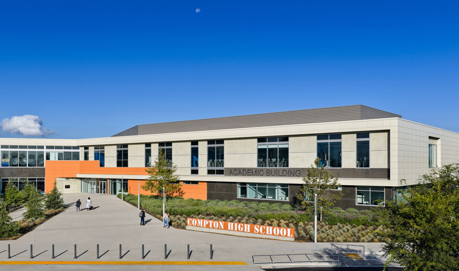 Front entrance of Compton High School; orange sign with the name, modern facade and greenery in front of blue skies