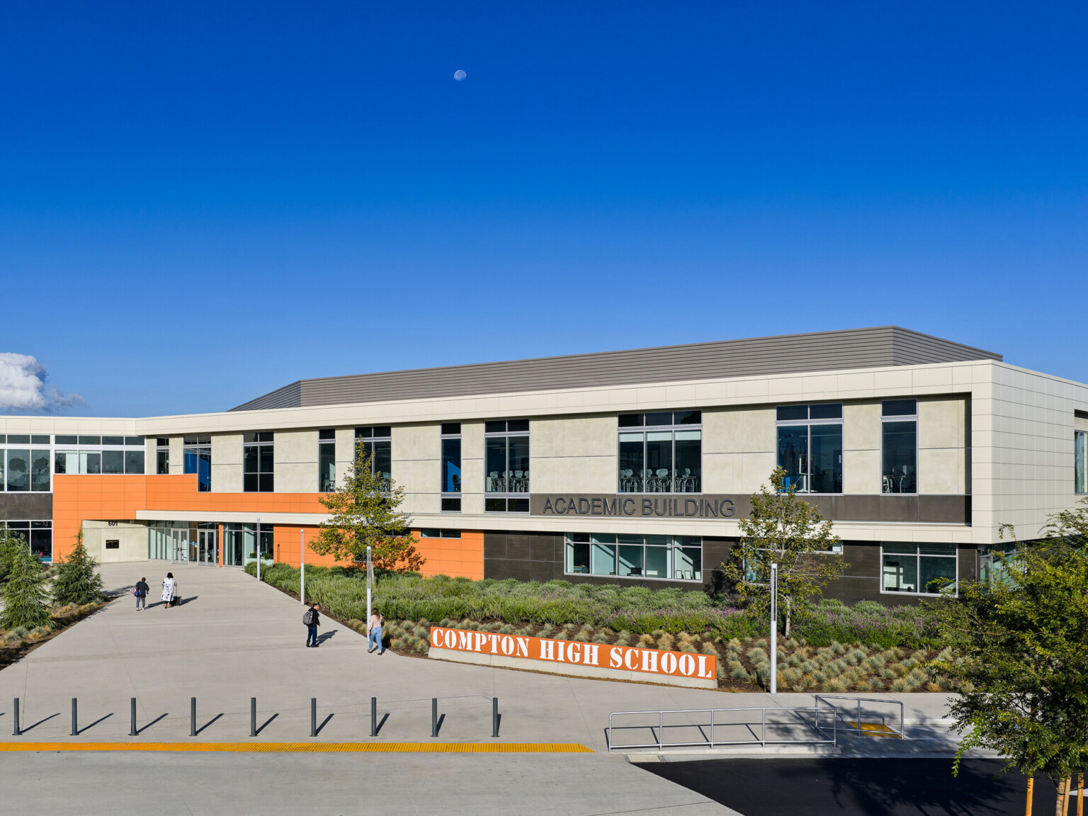 Front entrance of Compton High School; orange sign with the name, modern facade and greenery in front of blue skies