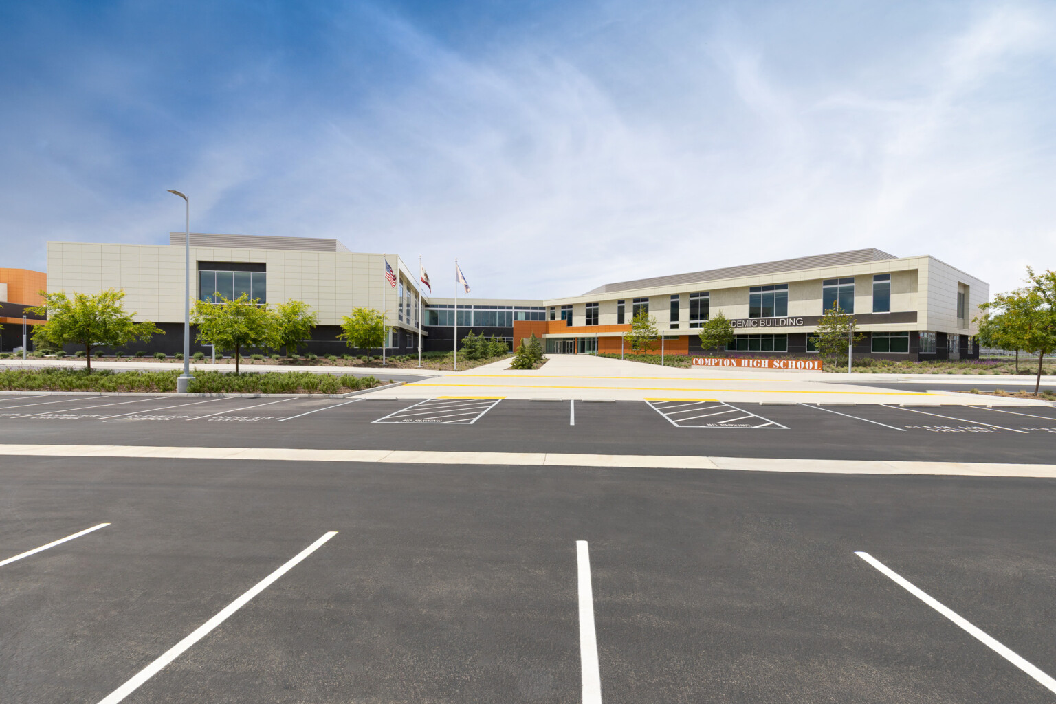 Front entrance of Compton High School; orange sign with the name, modern facade and greenery in front of blue skies
