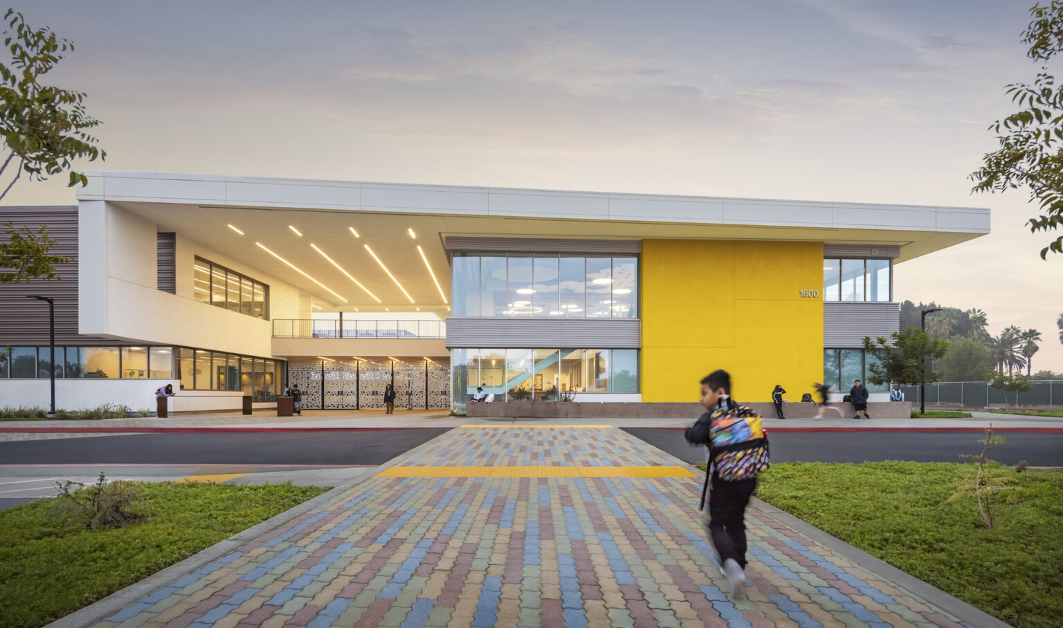 Front entrance of elementary school with multicolor stone path leading to yellow building with grey accents and white screen gates
