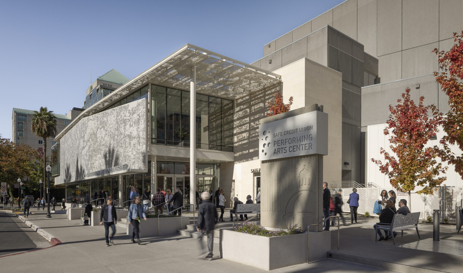 Outside a new performing arts center, facility sign displayed with SAFE Credit Union Performing Arts Center written, people gathered on benches and steps, large solar canopy hangs over patio