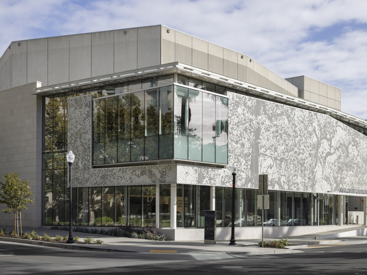 Corner view of a modernized brutalist building, corner glass room hangs over public patio, perforated metal façade provides shade, trees reflect on the glass, scrim