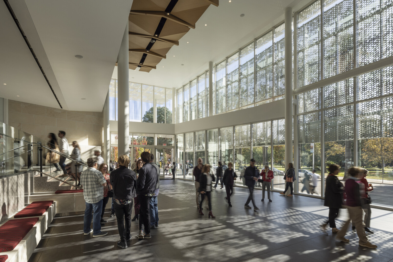 Two-story lobby space lit by floor to ceiling glass windows; wood sculpture covers ceiling; perforated light shines through the exterior lacy scrim, groups of theater enthusiasts gather