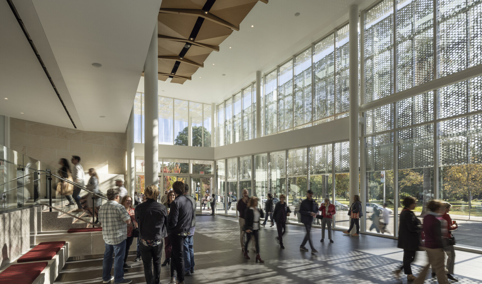 Two-story lobby space lit by floor to ceiling glass windows; wood sculpture covers ceiling; perforated light shines through the exterior lacy scrim, groups of theater enthusiasts gather