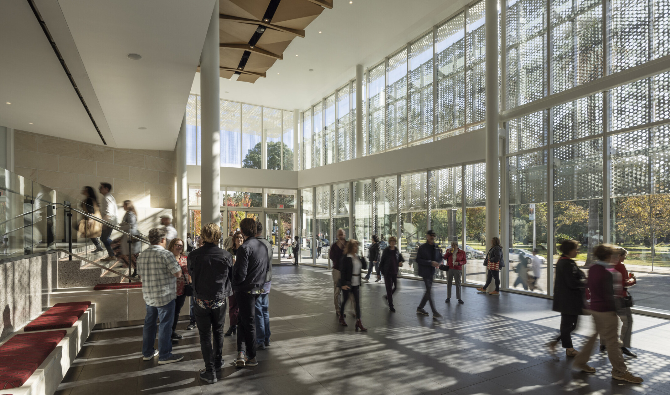 Two-story lobby space lit by floor to ceiling glass windows; wood sculpture covers ceiling; perforated light shines through the exterior lacy scrim, groups of theater enthusiasts gather