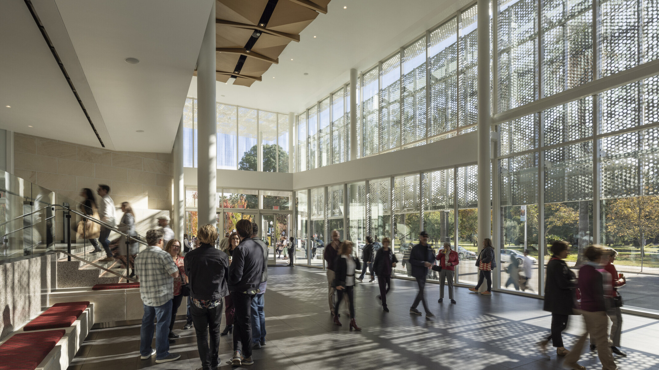 zTwo-story lobby space lit by floor to ceiling glass windows; wood sculpture covers ceiling; perforated light shines through the exterior lacy scrim, groups of theater enthusiasts gather