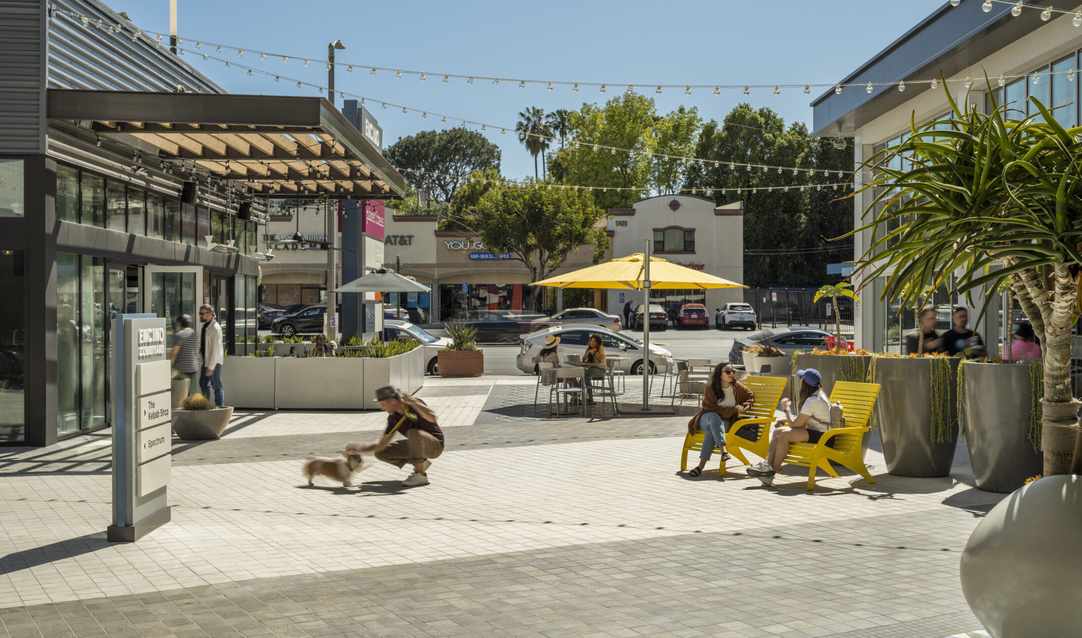 bright lively space in between retail storefronts; people sitting at tables and chairs under yellow umbrellas; a person engaging with a dog in the front