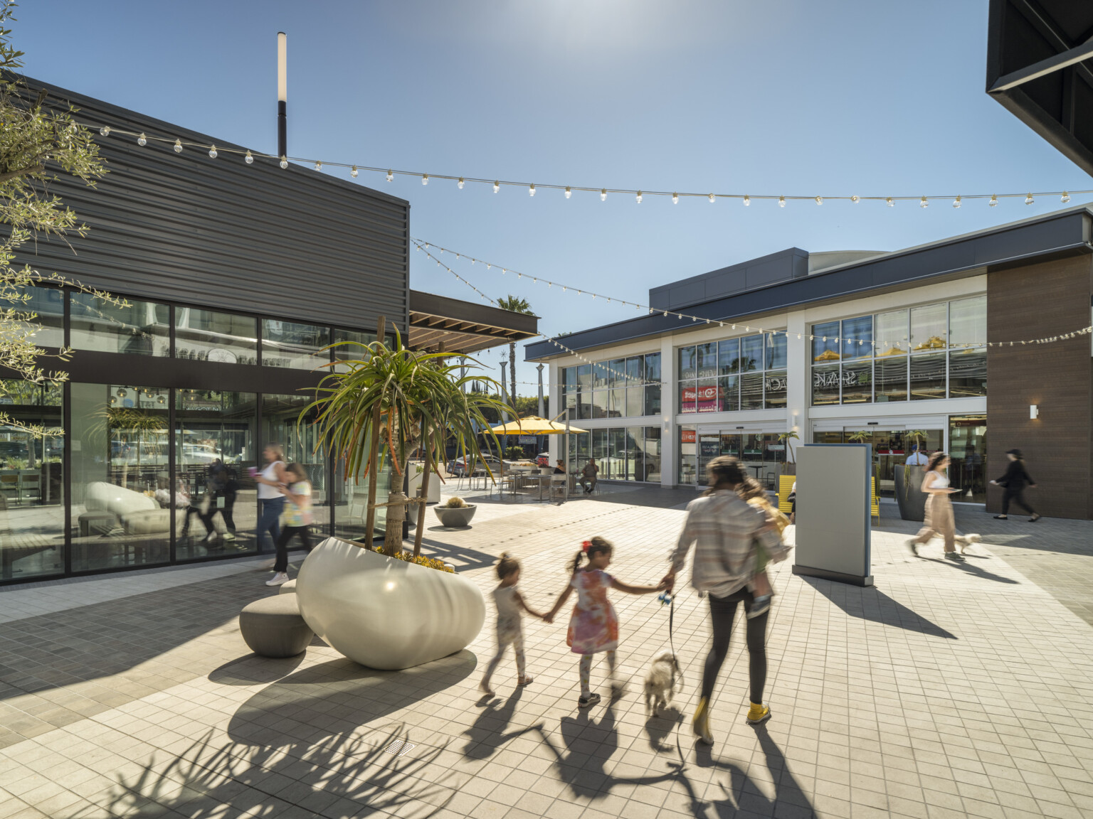 Modern outdoor shopping area with buildings, a family walking on a paved walkway with light-colored tiles with string lights overhead