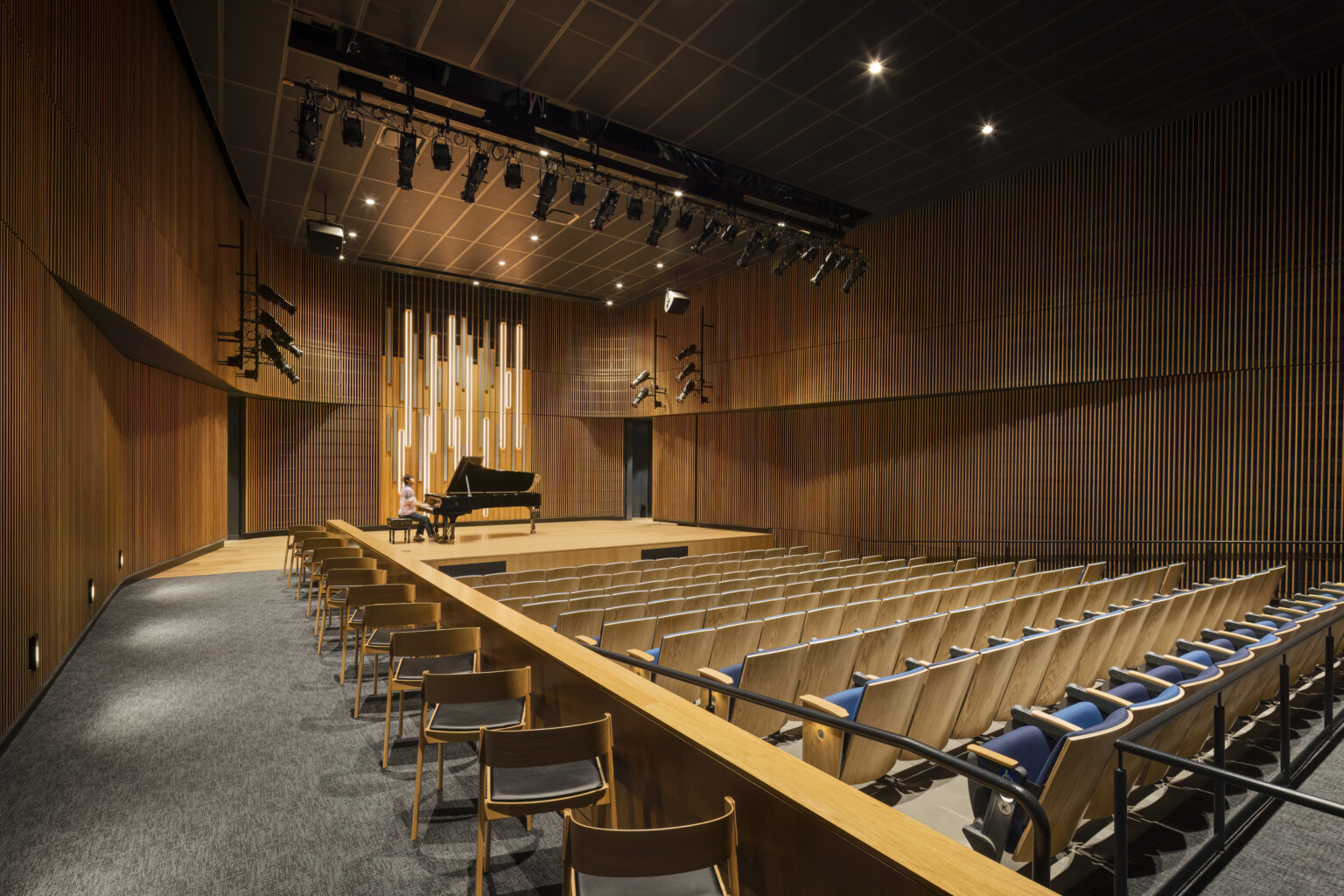 Concert hall in the Irvine Valley College fine arts building; large wood acoustic room with chairs facing the elevated stage; performer on a piano on the stage; lighting on the performer