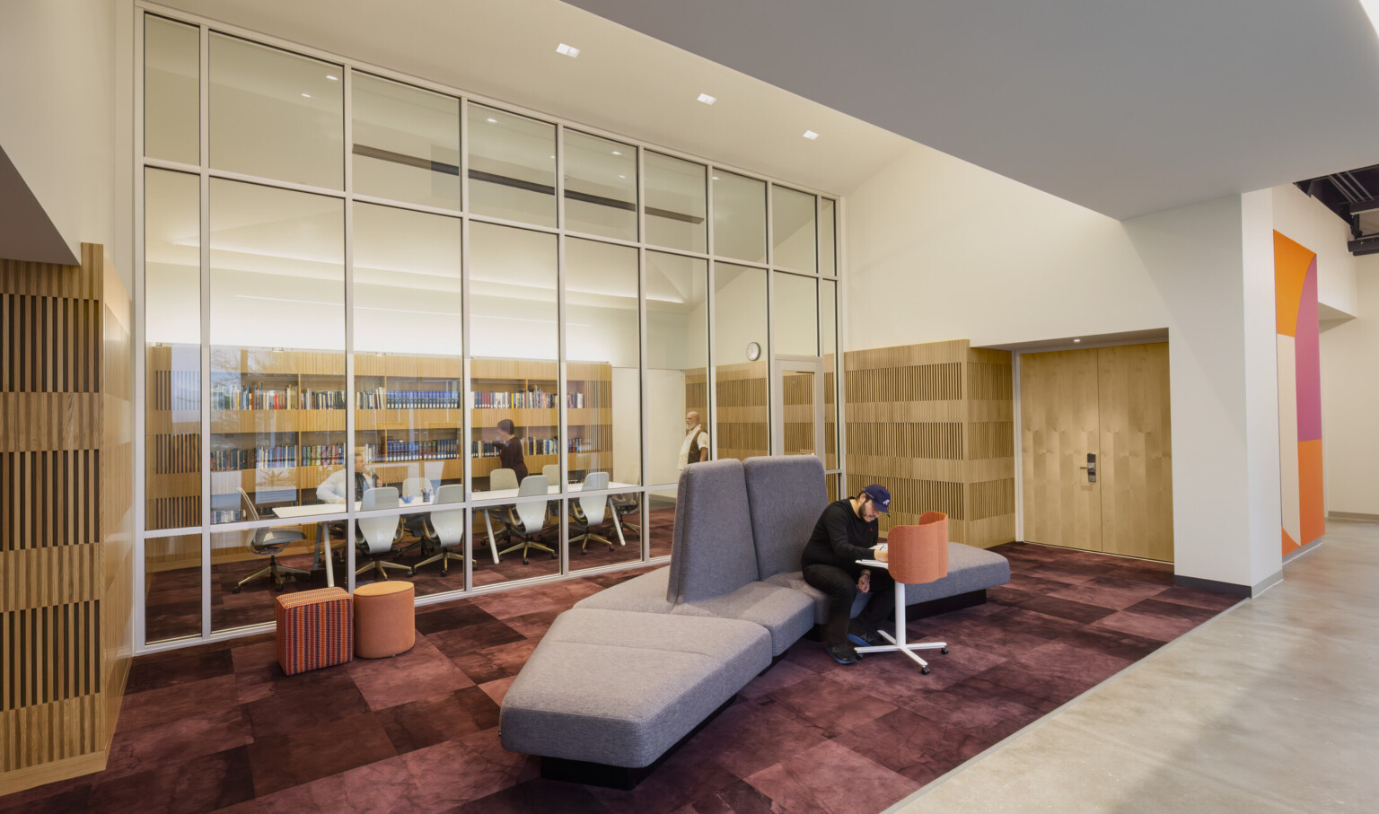 study space in the Irvine Valley College Fine Arts Building; conference space in the background separated by glass; comfortable seating in front of the glass; maroon carpet with orange poufs and a large gray lounge couch