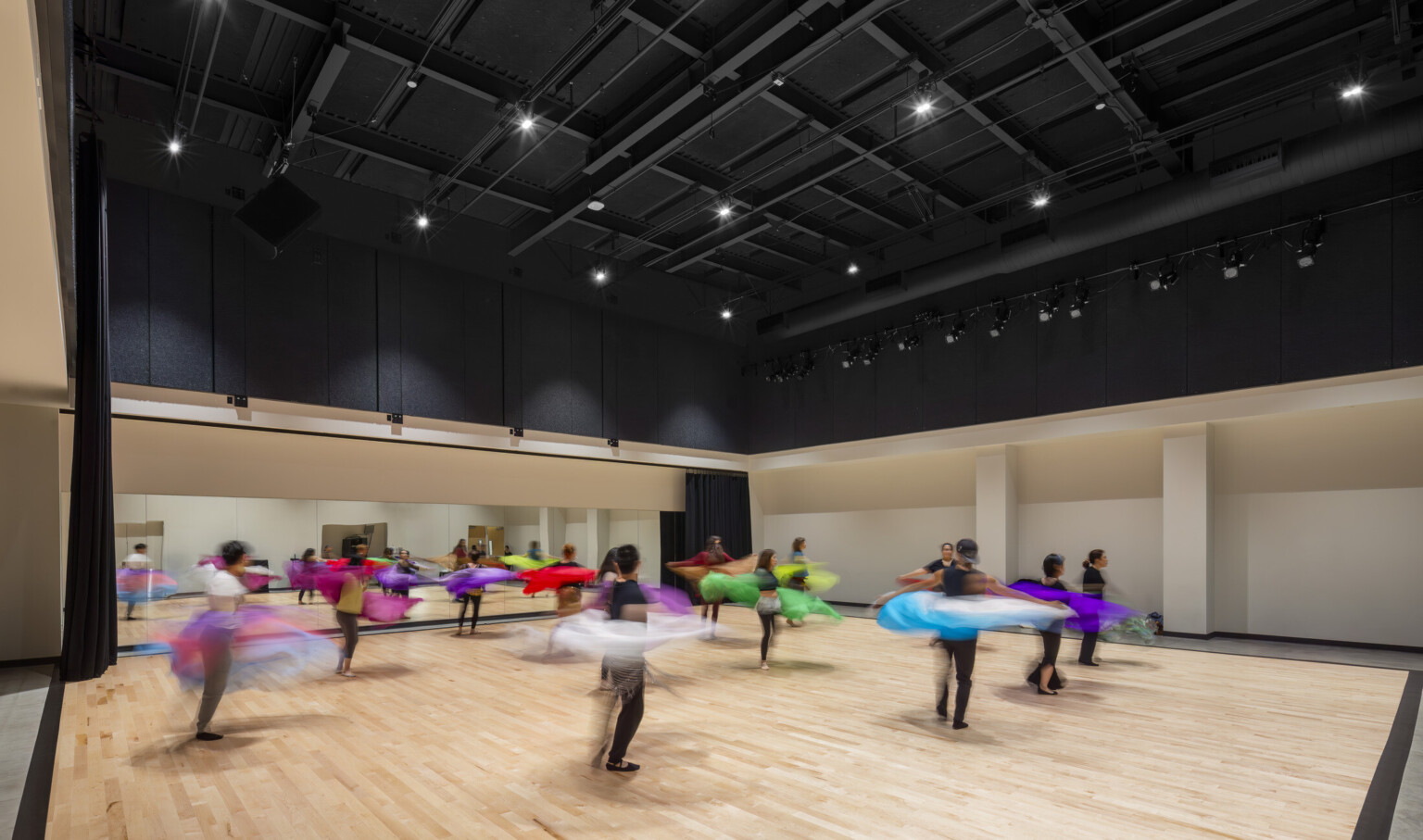 Dance hall at Irvine Valley College Fine Arts Building' light wood dance floor with dark ceiling and spotlights on dancers twirling around in bright colored fabric; mirror and barre at the back wall