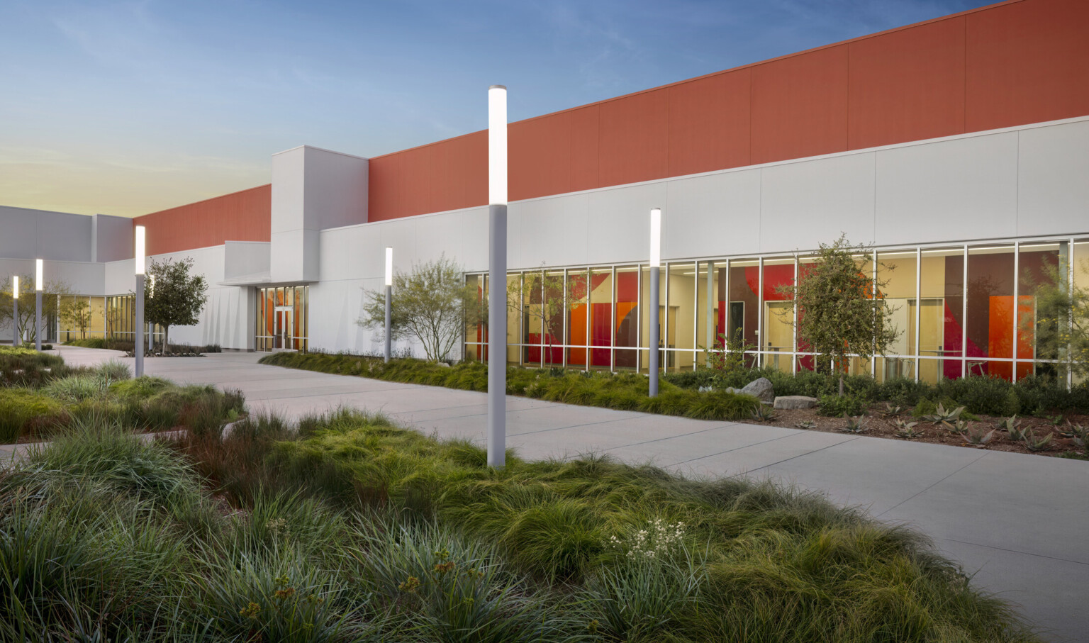 entrance of the Irvine Valley College Fine Arts Building at night; grass flanks the walkway to the modern white and orange building; floor to ceiling windows peek into the colorful interior