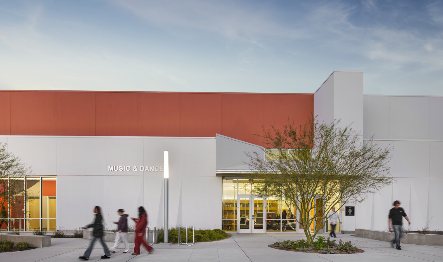 Front entrance of Irvine Valley College Fine Arts Building; modern white and orange building with seating and green elements in front; students walking to and from class