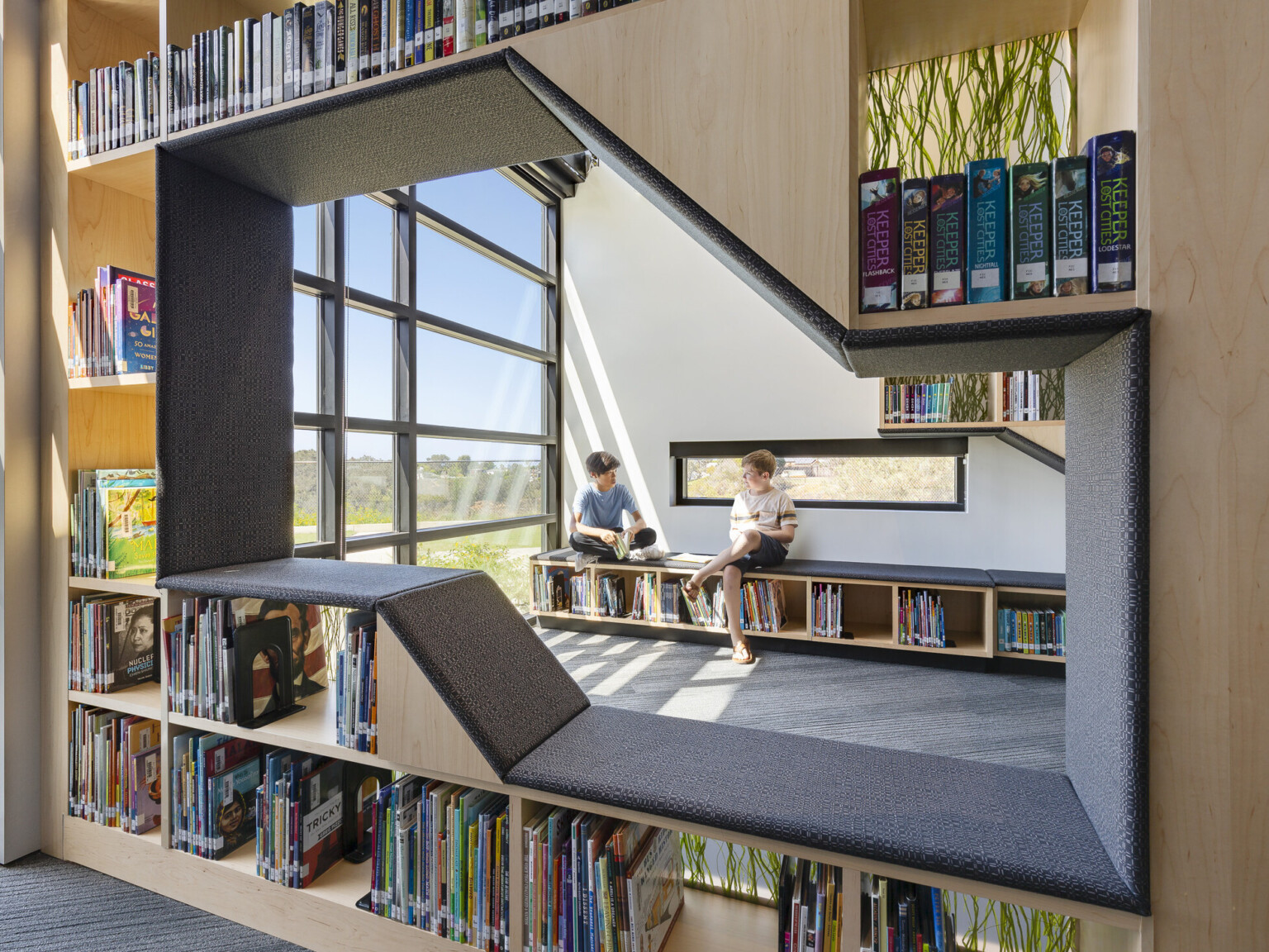 library of Del Mar Heights School; indoor outdoor learning space with large floor to ceiling windows; coastal community design with sand and wood tones with greenery; children reading in shelves with benches build into them library of Del Mar Heights School; indoor outdoor learning space with large floor to ceiling windows; coastal community design with sand and wood tones with greenery; children reading in shelves with benches build into them