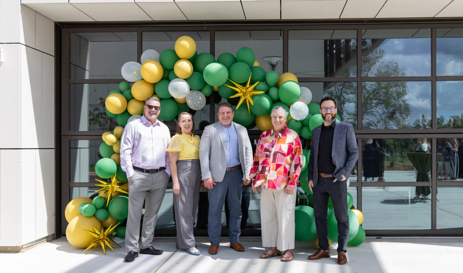 Five individuals stand in front of a large, windowed building entrance decorated with a balloon arch made of green, yellow, and white balloons, along with gold star-shaped decorations.