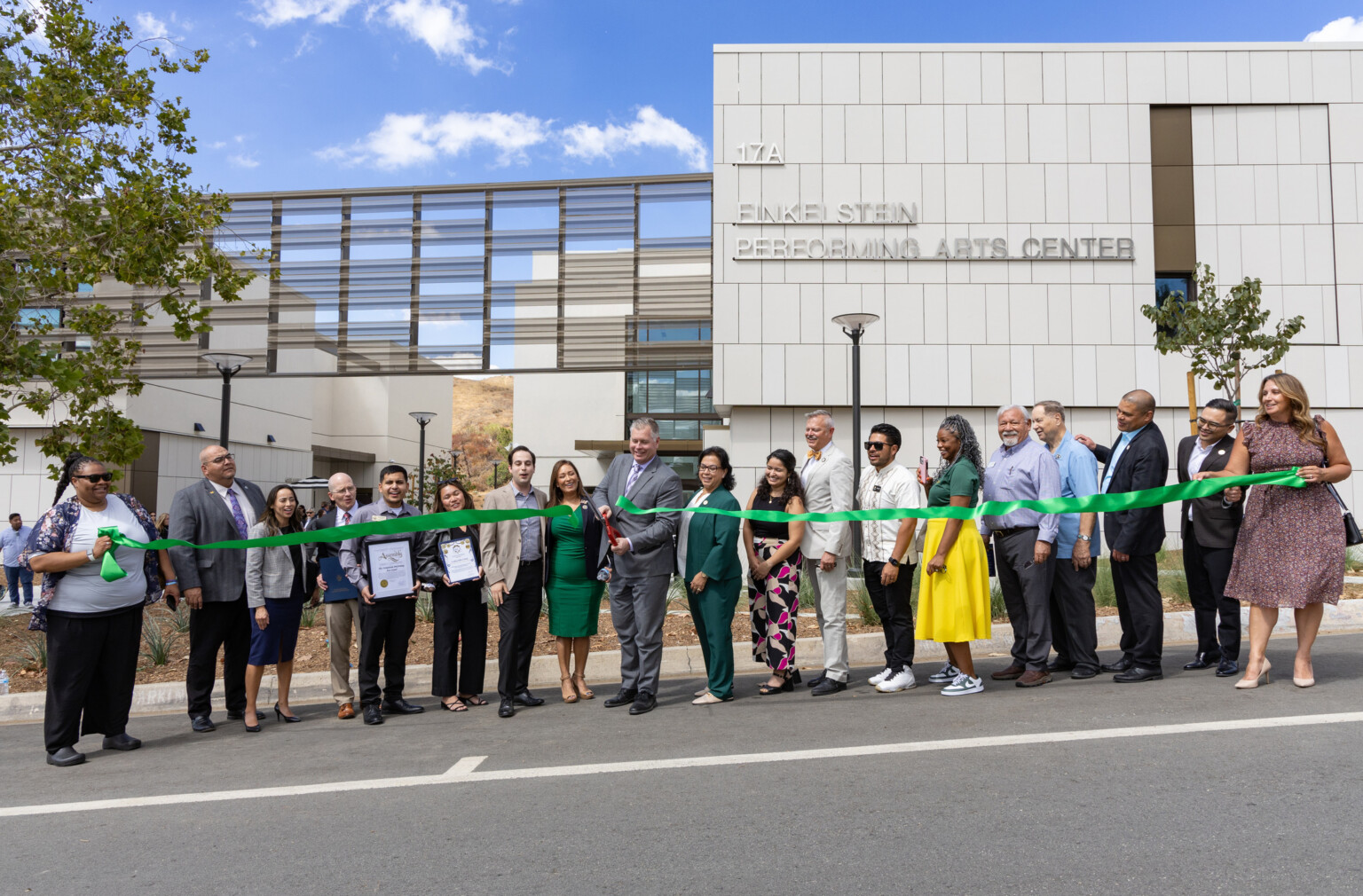 A group of people stands in front of a modern building. They are holding a long green ribbon, which is being cut by a person in the center using large scissors.