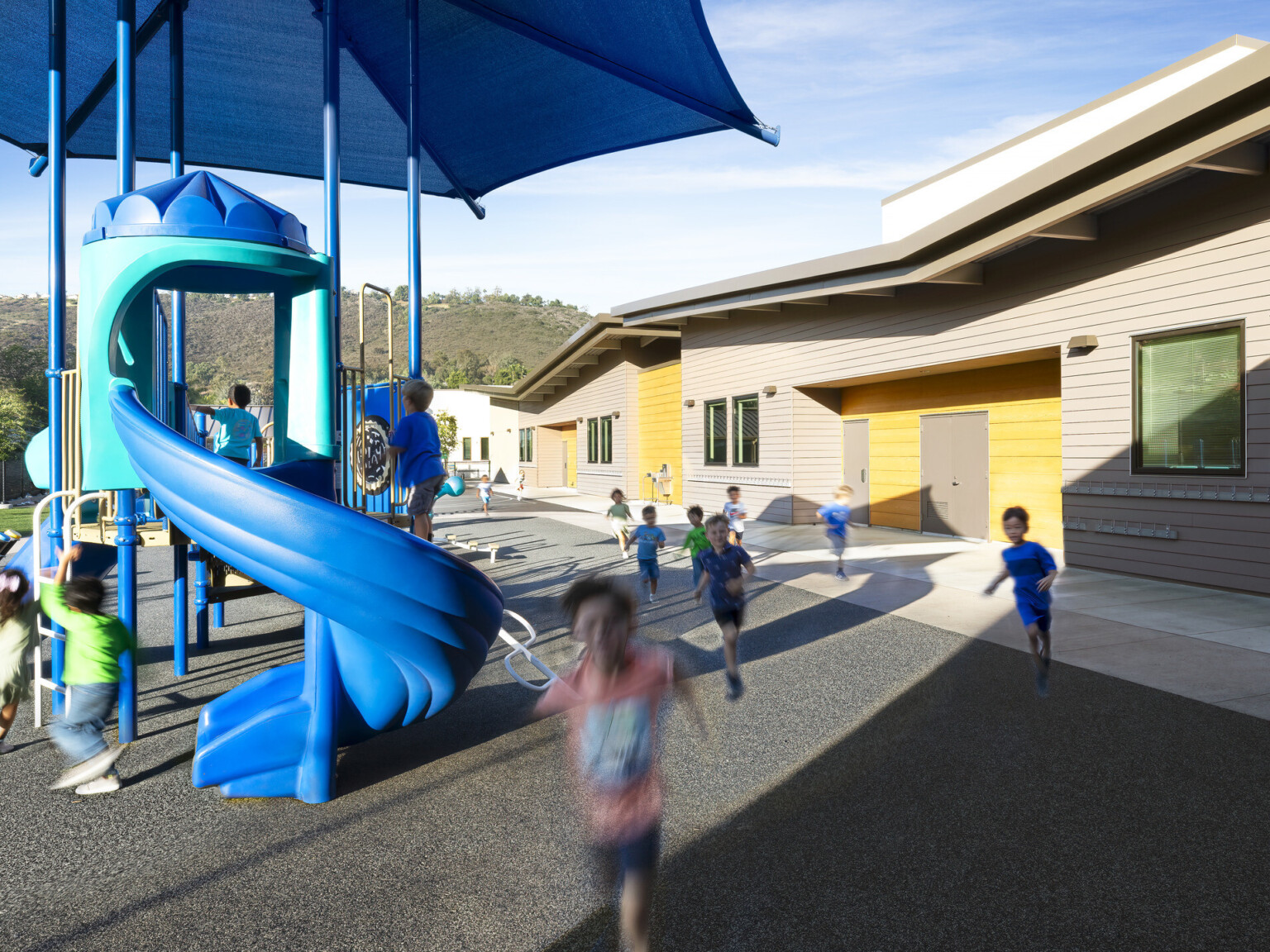 Single story angular roof building next to elementary school playground with blue canopied slide