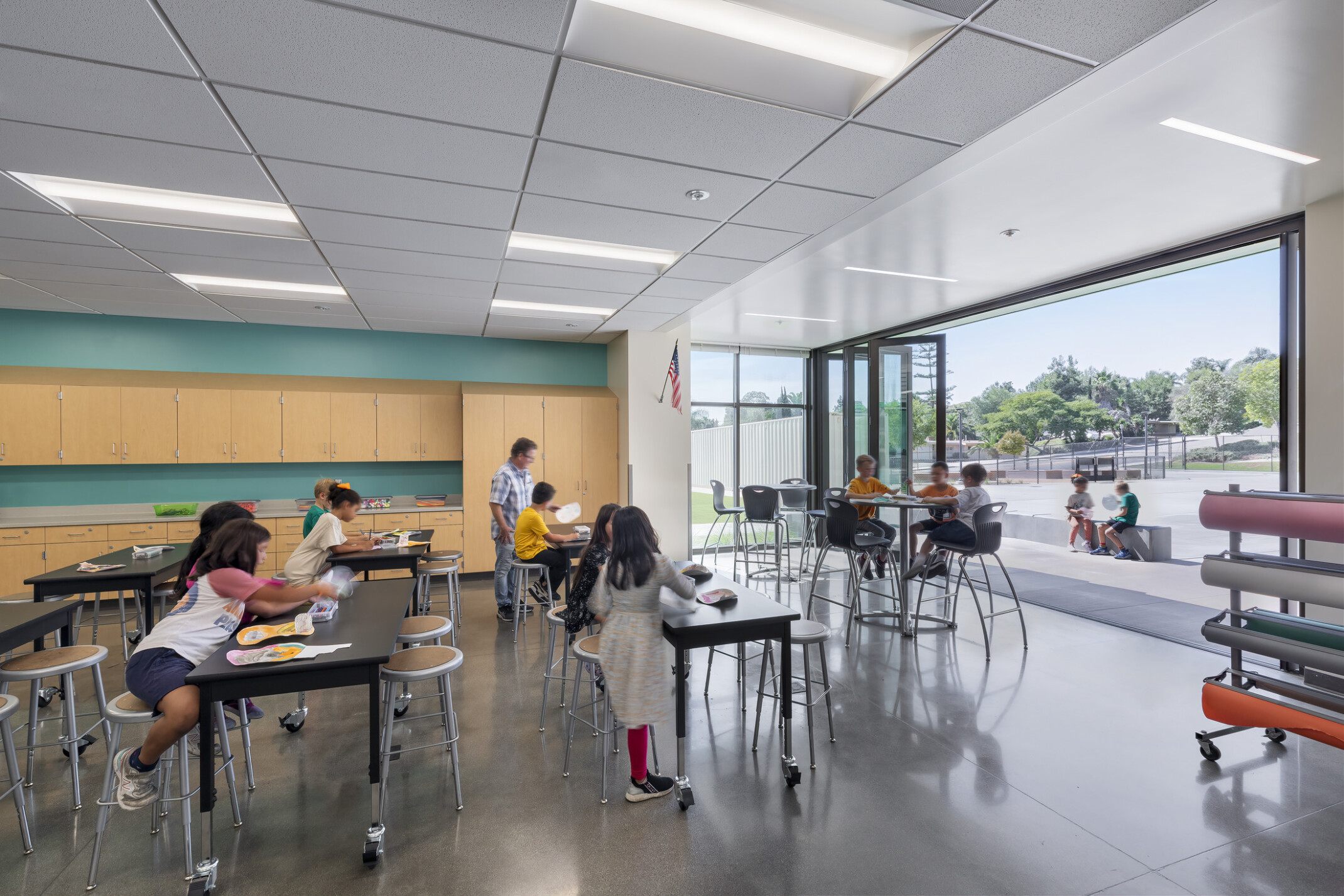 Richland Elementary School learning space with wood cabinets and mixed height seating spaces; floor to ceiling window wall opens to playground for campus design flow