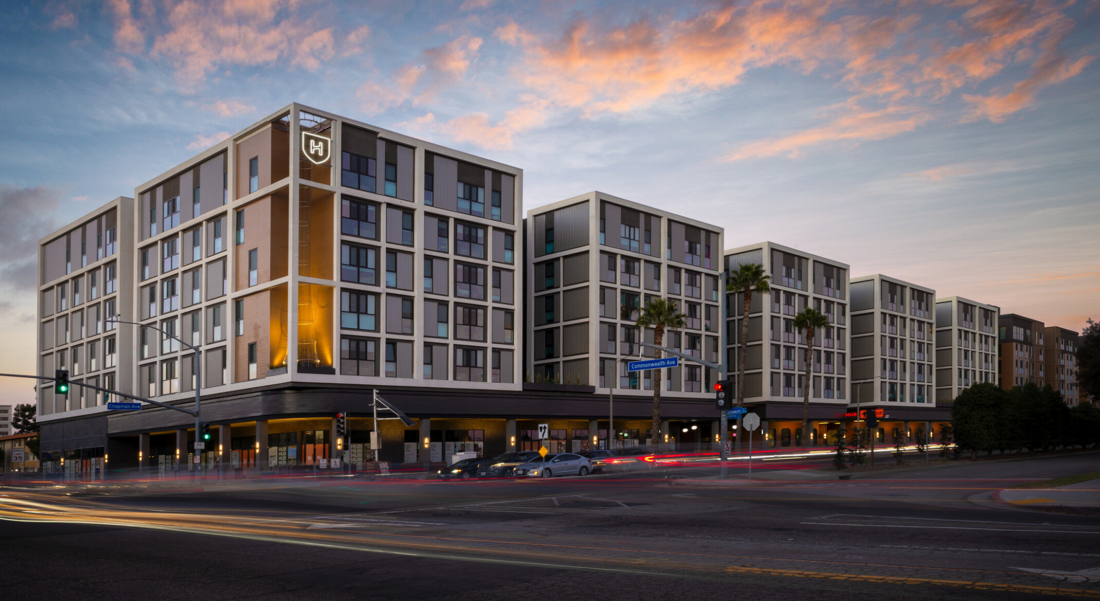 View of Hub Fullerton's campus gateway corner, with ground-floor retail and street traffic in the foreground. The façade steps across multiple volumes, transitioning from one material palette to another.
