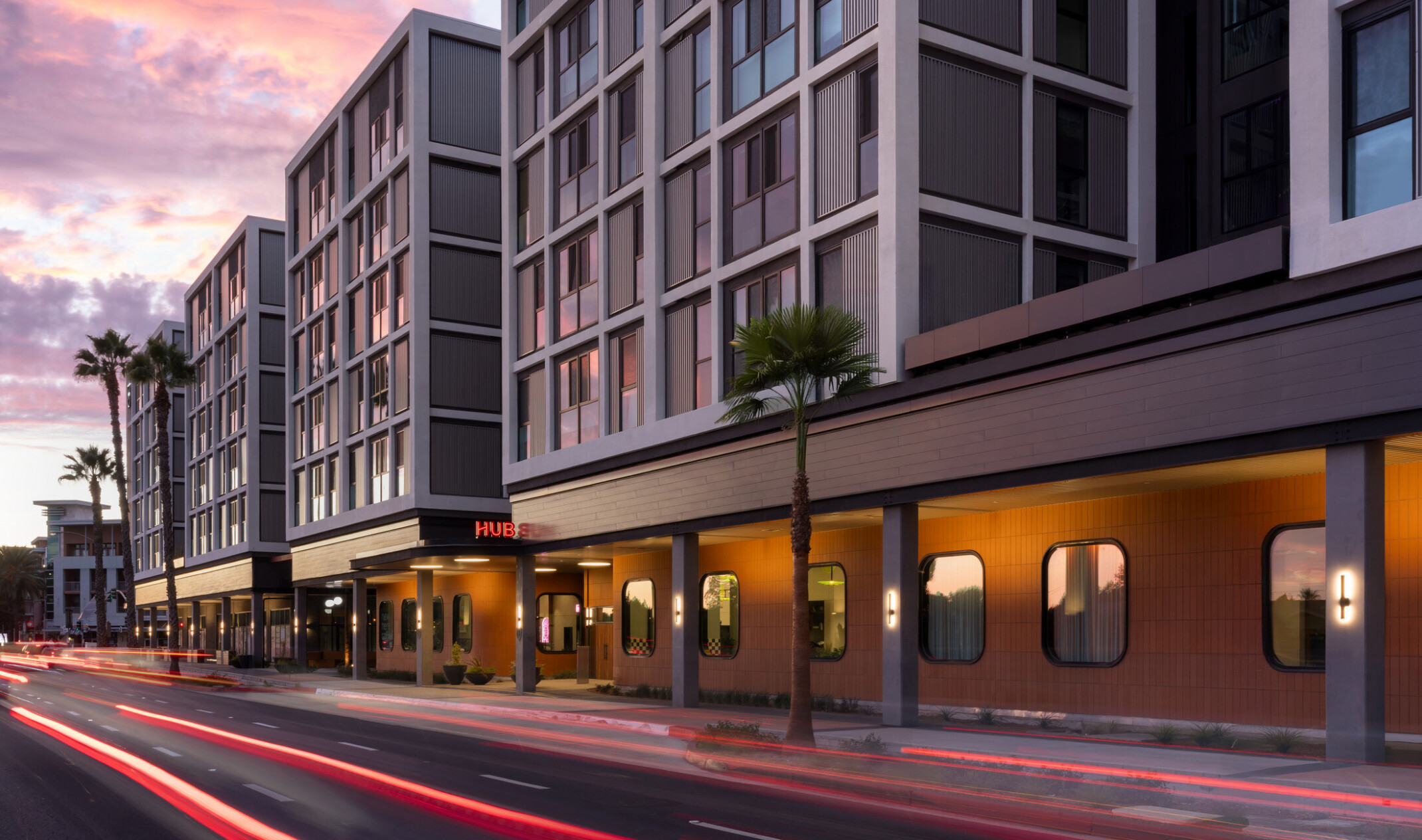 Hub Fullerton's main entrance at sunset, with an illuminated canopy and Hub logo, light trails from passing cars, and lighting glowing beneath the building's projecting upper edge.