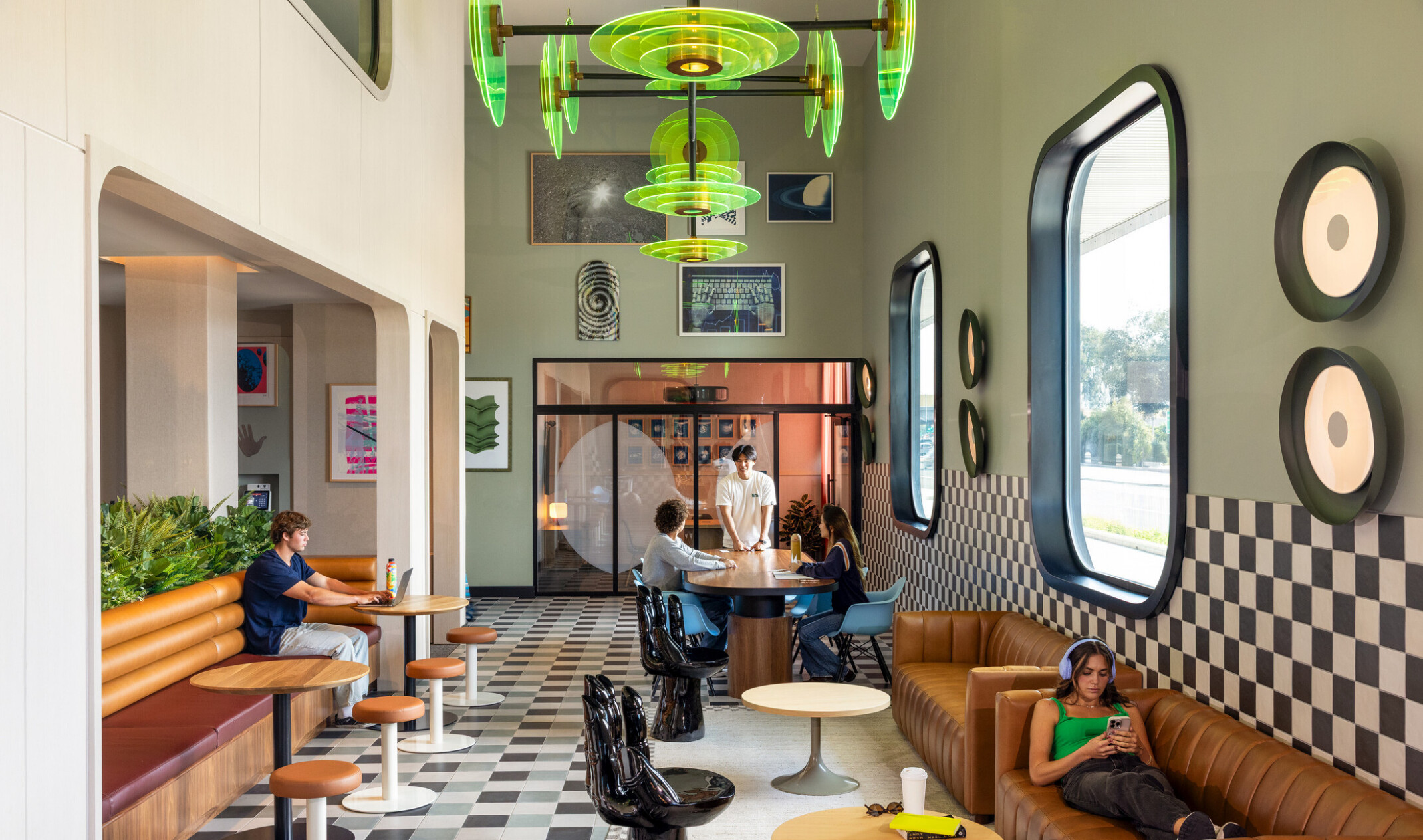 Interior view of Hub Fullerton's ground floor lounge, with an aesthetic featuring black hand-shaped chairs, a communal table, booths, framed artwork on the walls, and a neon-green acrylic light fixture. Students are gathered throughout the space.