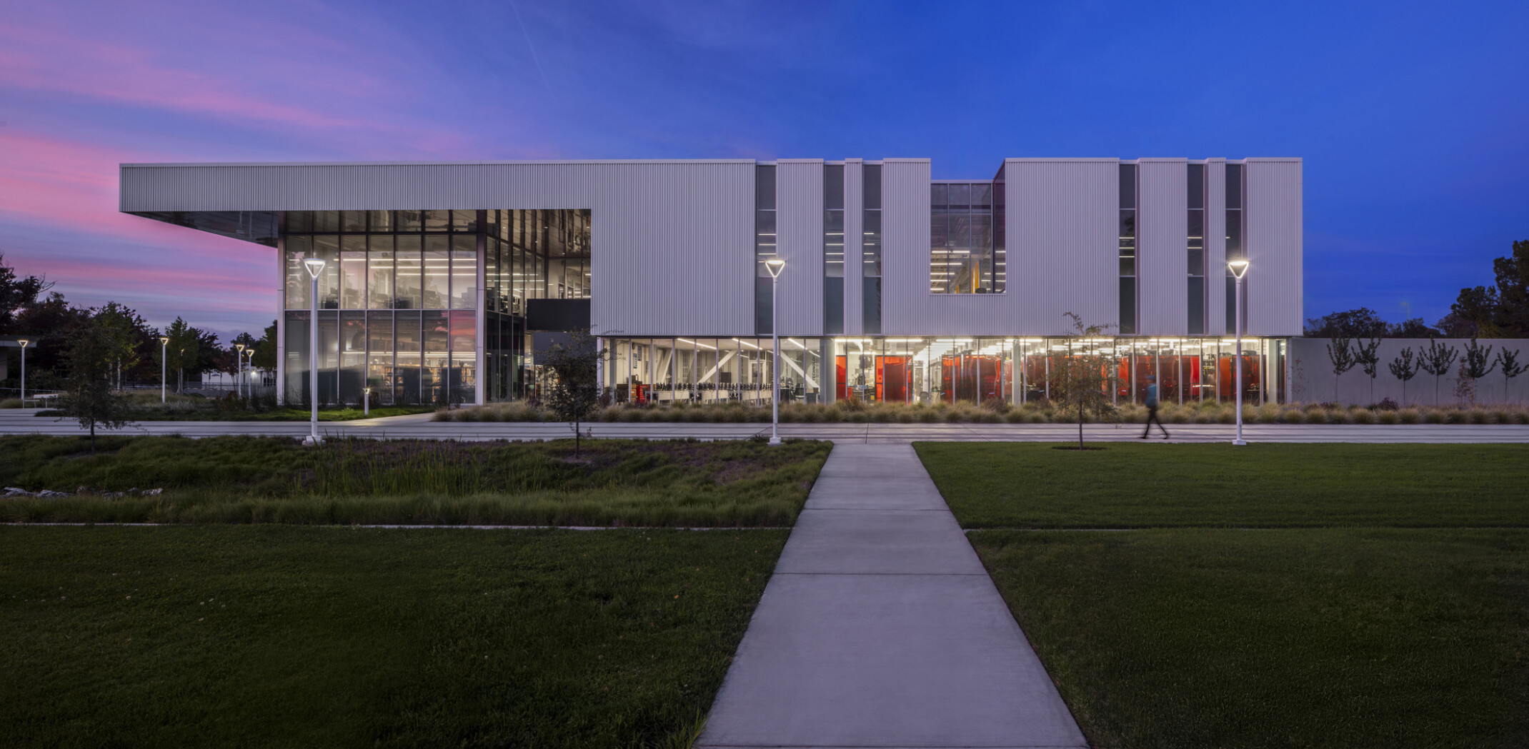 evening view of a modern, rectangular building; white facade with recessed ground level and entrance glass walls