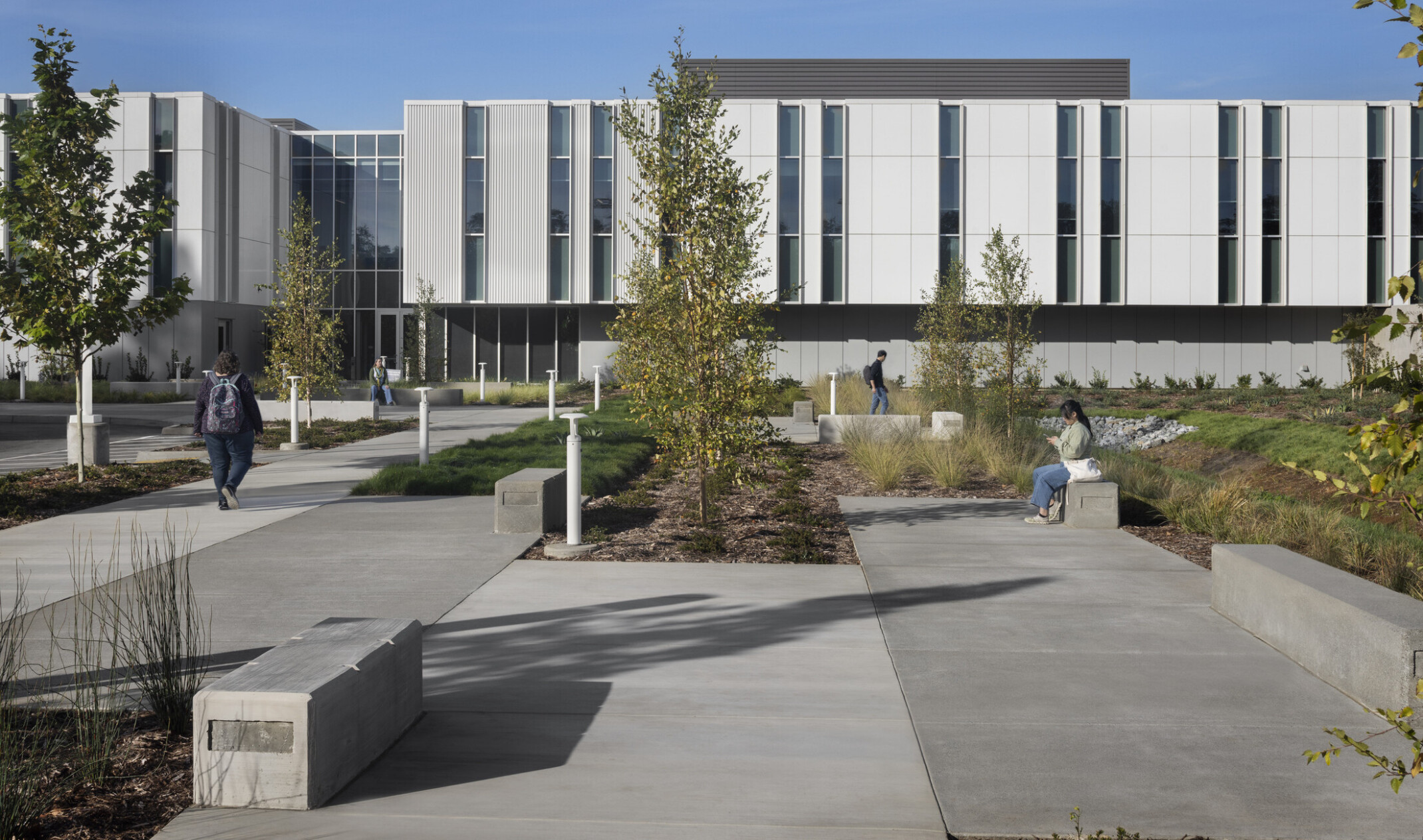 view of a modern, rectangular building; white facade with recessed ground level and entrance glass walls; foreground sitting space and greenery