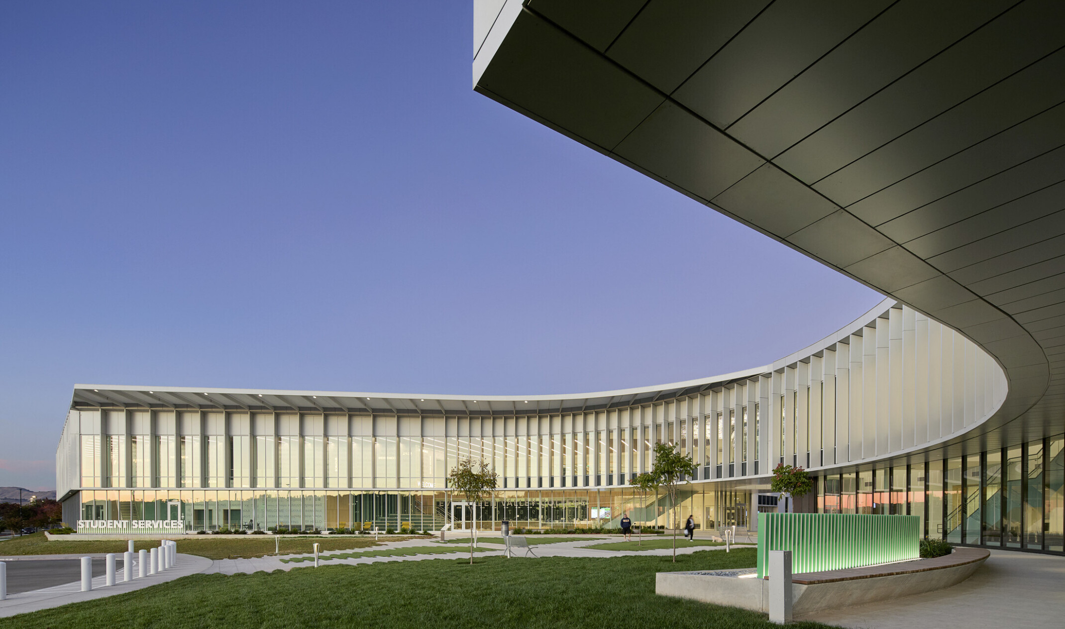 modern curved building with open green space and walking area beneath building; curved glass facade against a blue sky