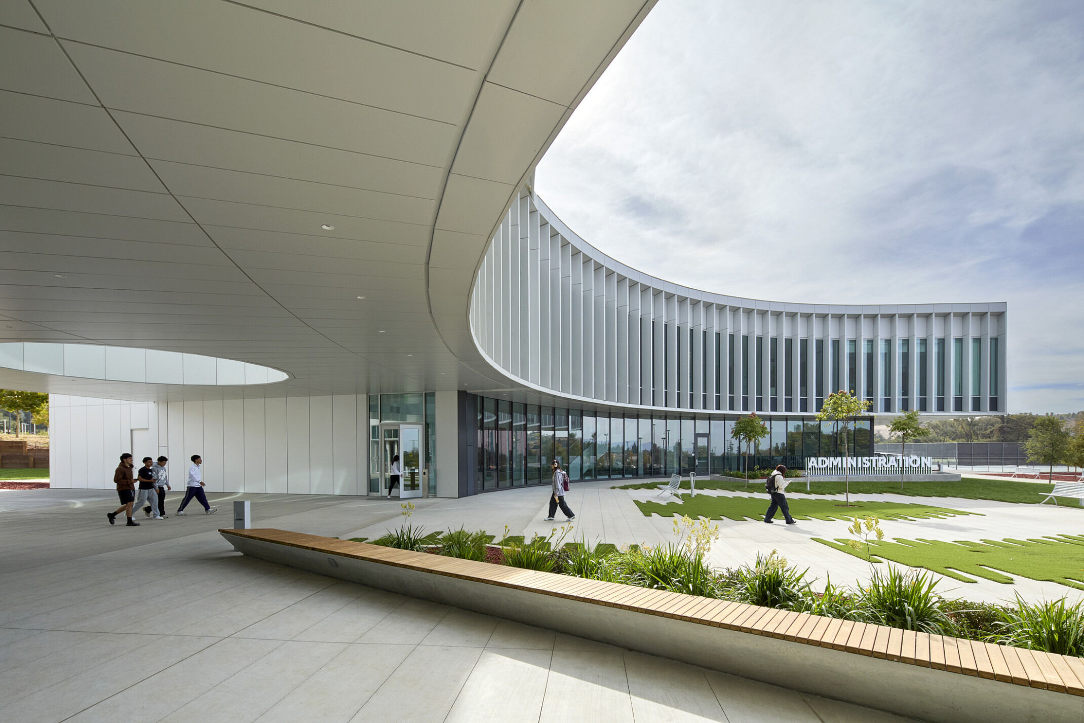 modern curved building with open green space and walking area beneath building; curved glass facade against a blue sky