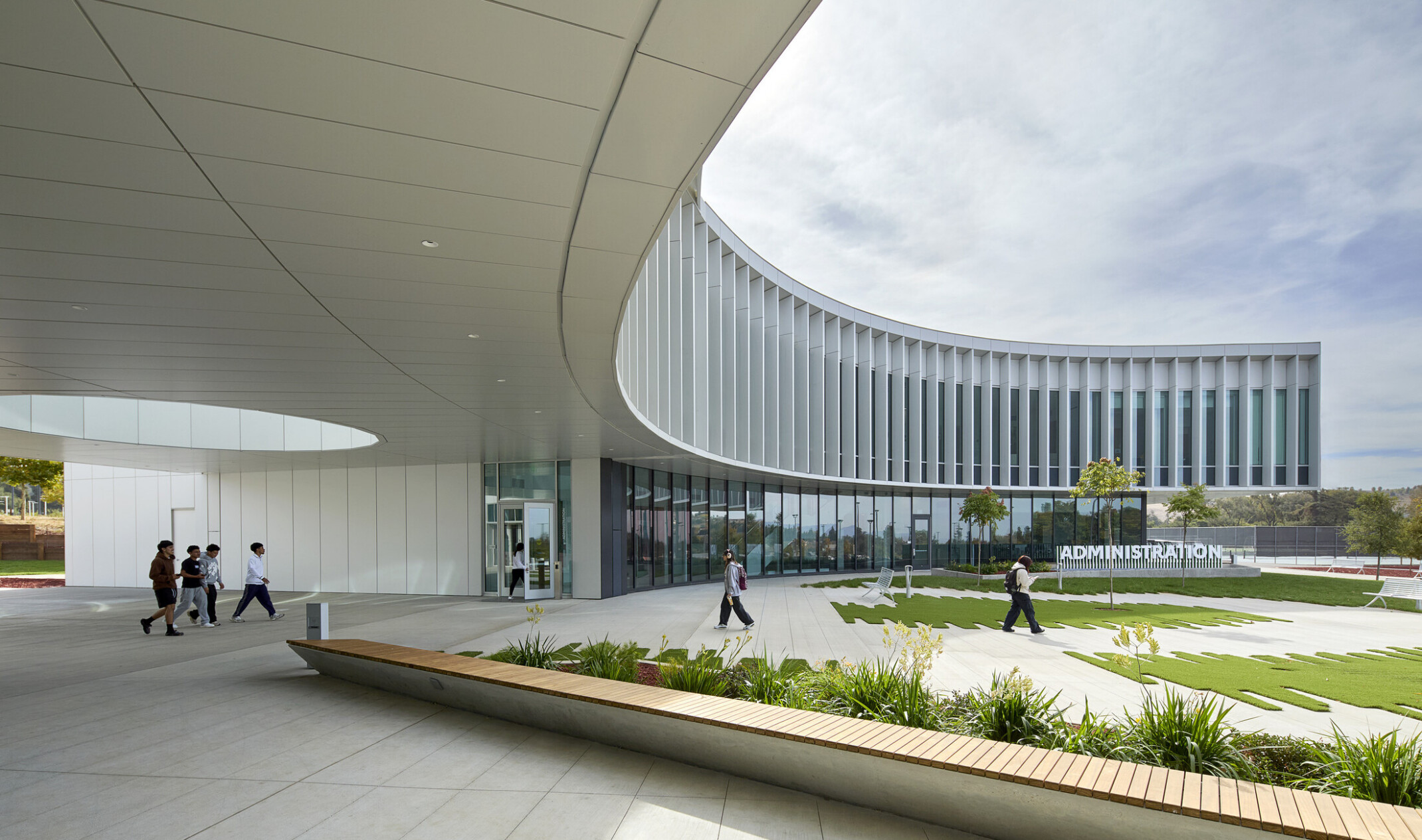modern curved building with open green space and walking area beneath building; curved glass facade against a blue sky