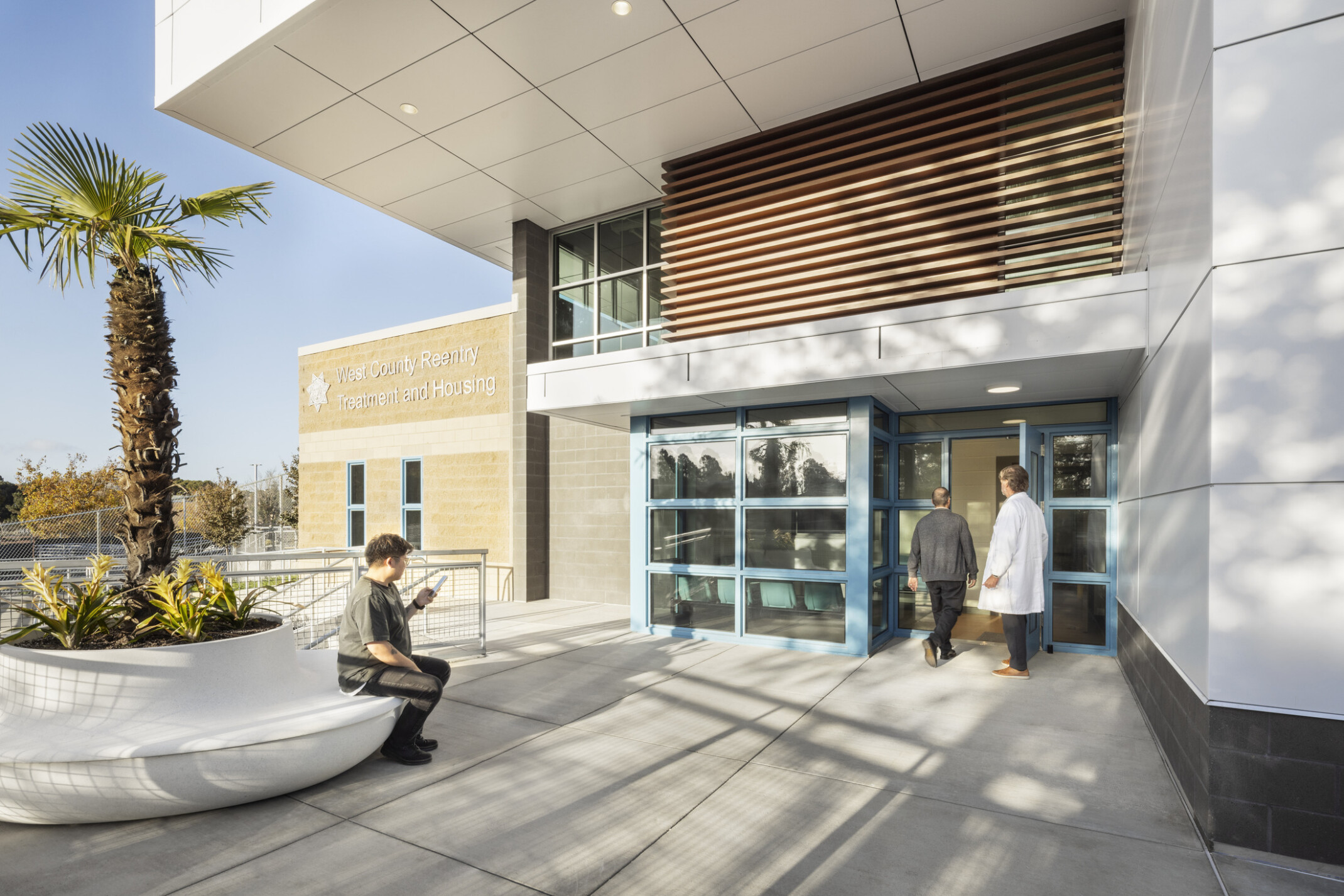 Entrance for public, staff, and visitors accented with light brick, white panels, blue-accented windows, and a palm tree in the front.