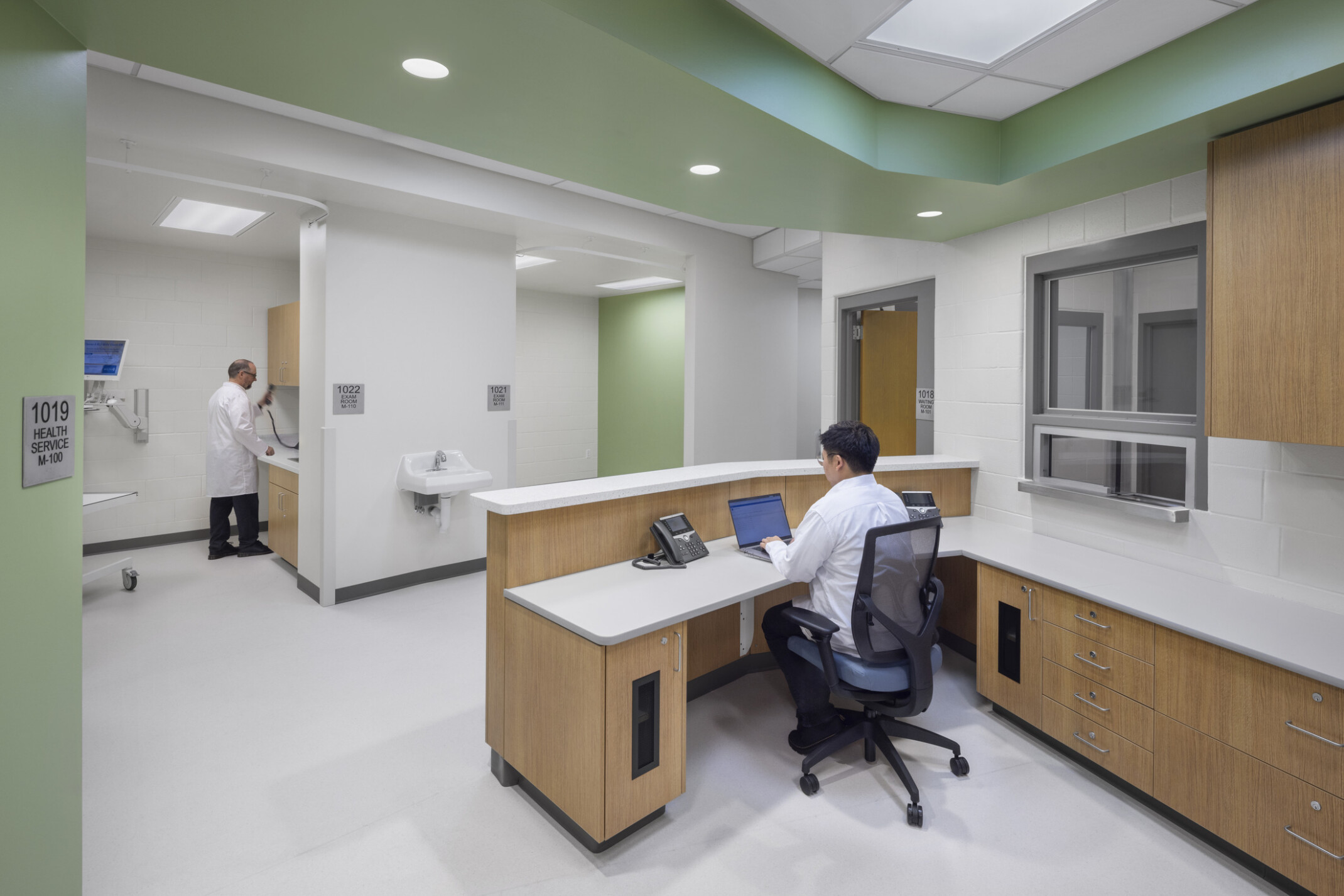 Medical Services exam rooms with a person at a desk in the foreground. Green walls, white floors, and light wood accent the space.