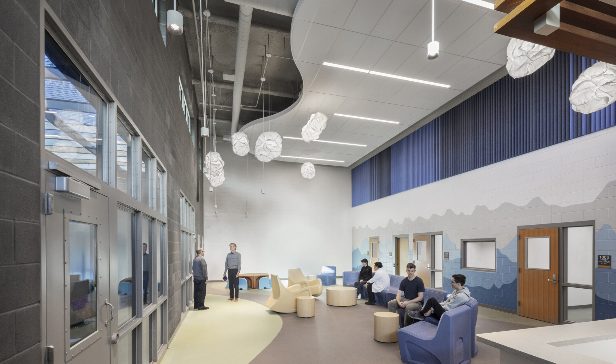 A visitation room with a serpentine acoustic ceiling; people sit on chairs and stools with a blue mountain mural behind them.