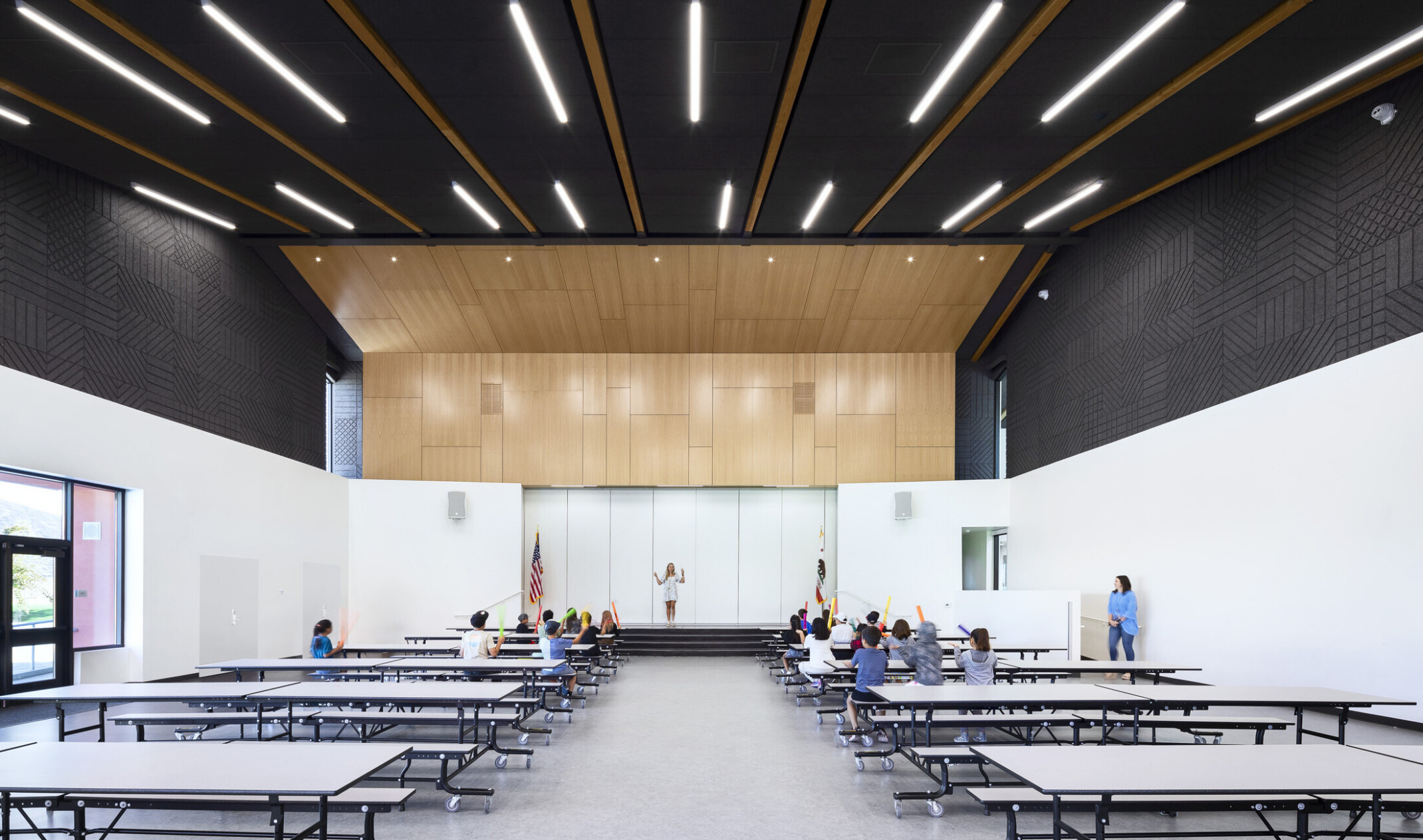 interior school performance space; long cafeteria tables face a small stage area with a white back drop
