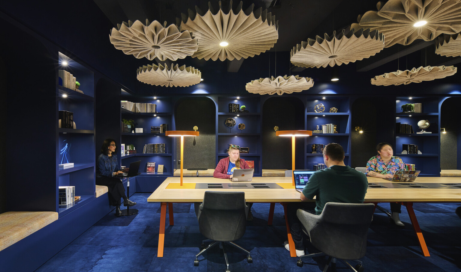 people working at a light wood table in a room with dark blue walls and ceilings, dark blue carpet and light wood chandeliers