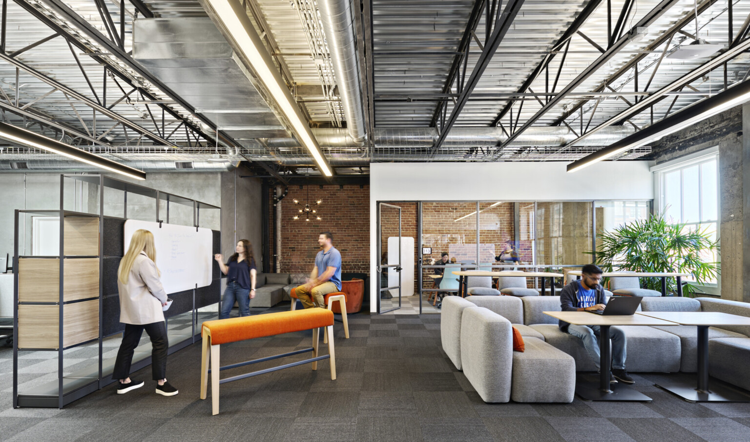Office common space with mixed seating, benches and chairs. Flexible shelf wall with attached white board. Exposed brick, industrial ceiling