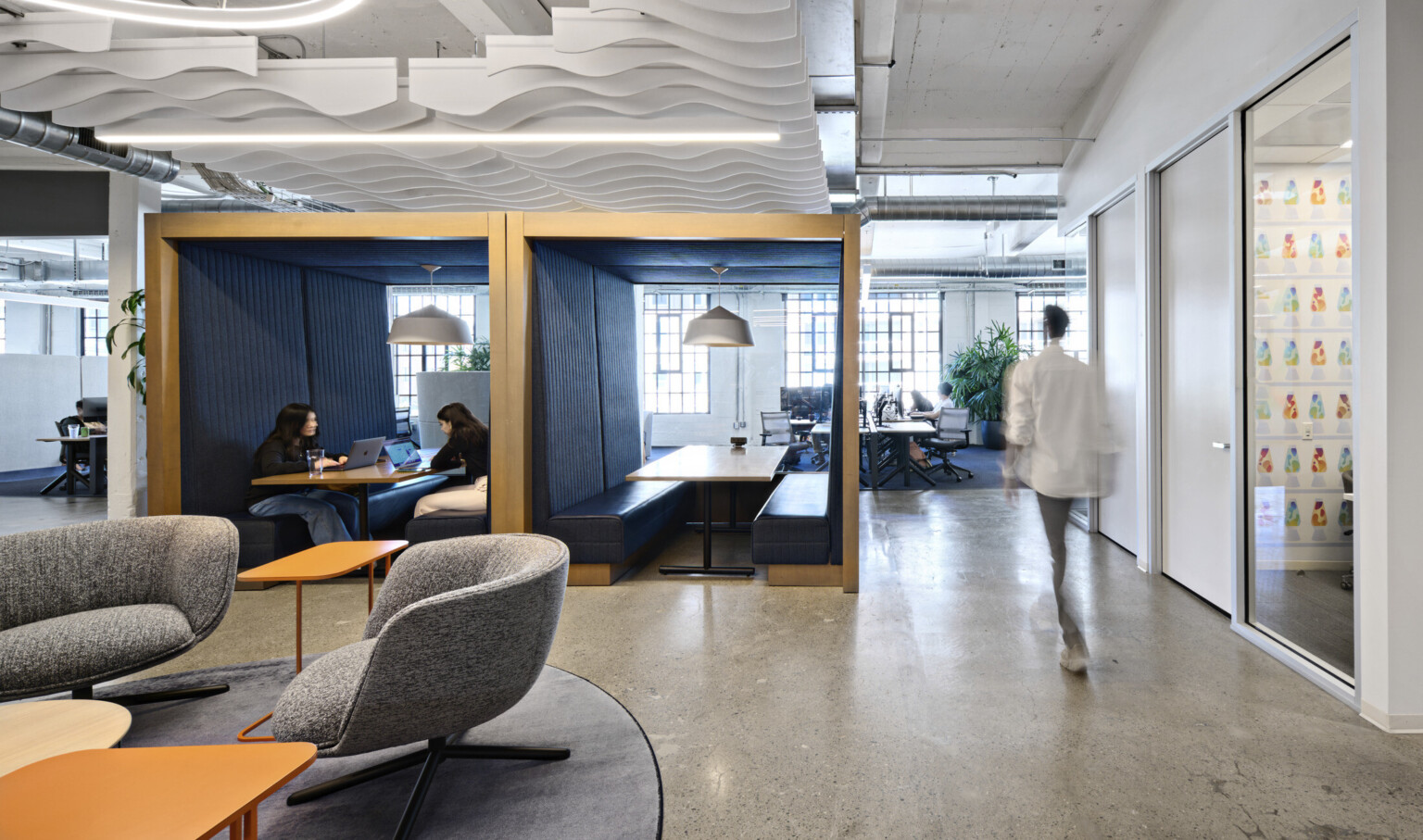 Office common space with grey armchairs and orange tables. 2 wood booths with bench seating at center enclosed front, back, above