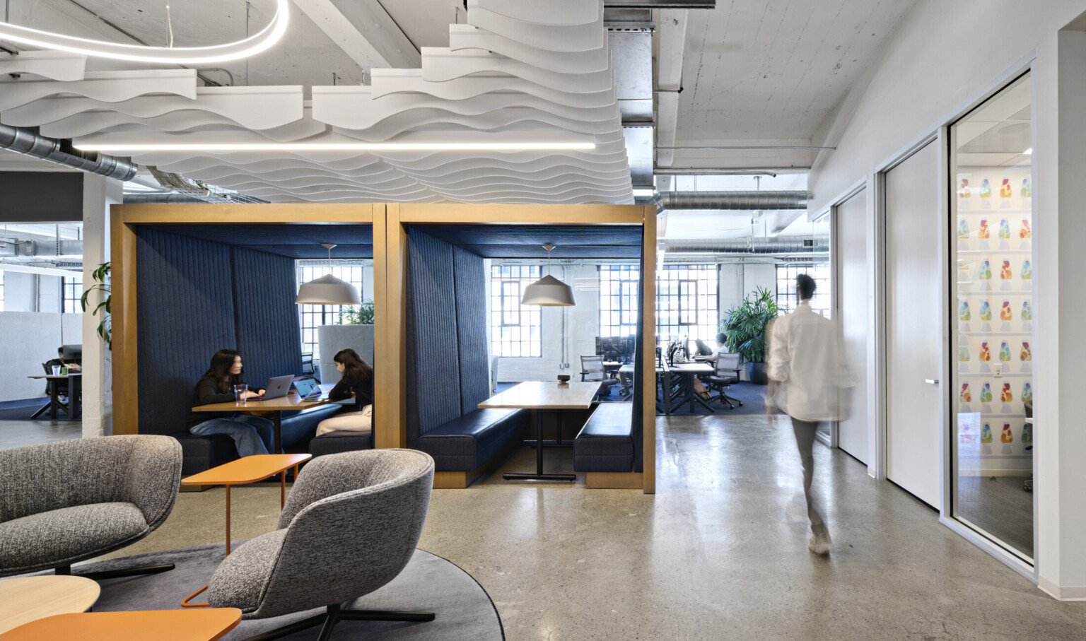 Office common space with grey armchairs and orange tables. 2 wood booths with bench seating at center enclosed front, back, above