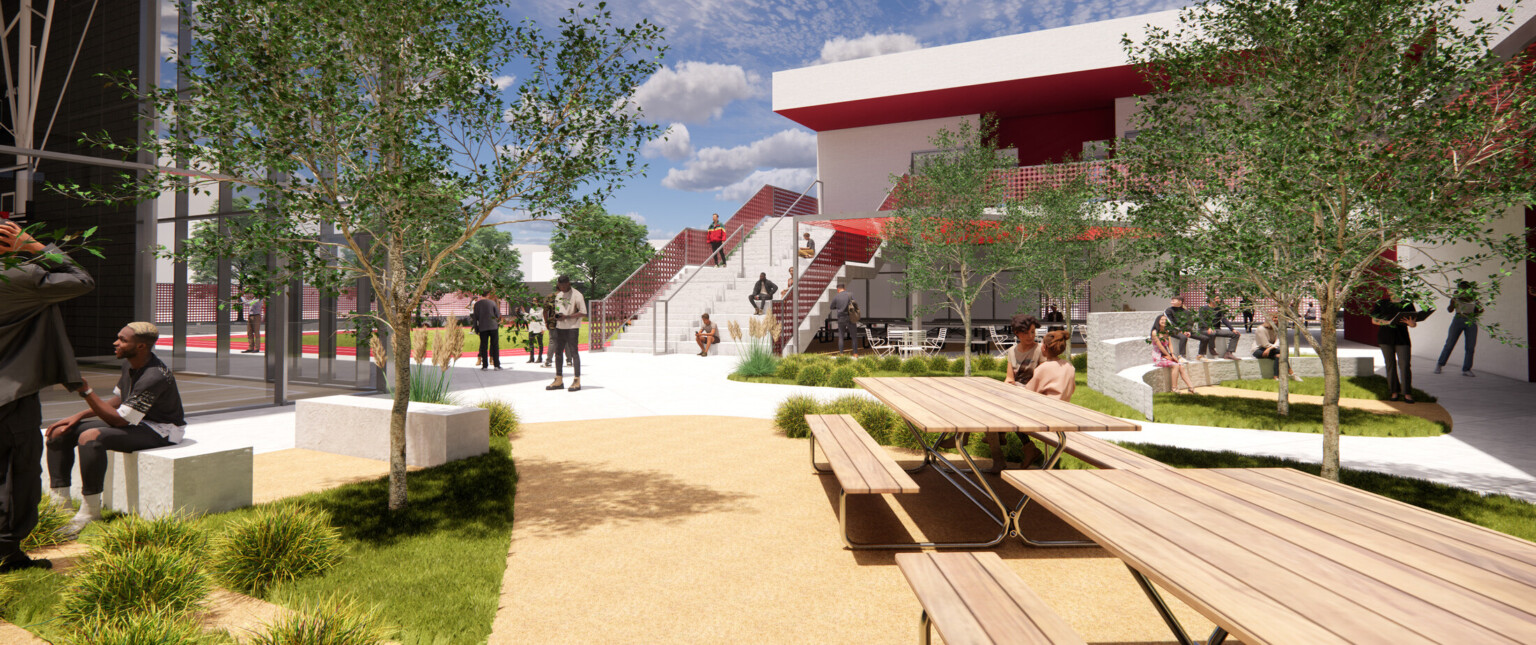 Outdoor courtyard with wooden picnic tables, lush landscaping, red accents, and people collaborating