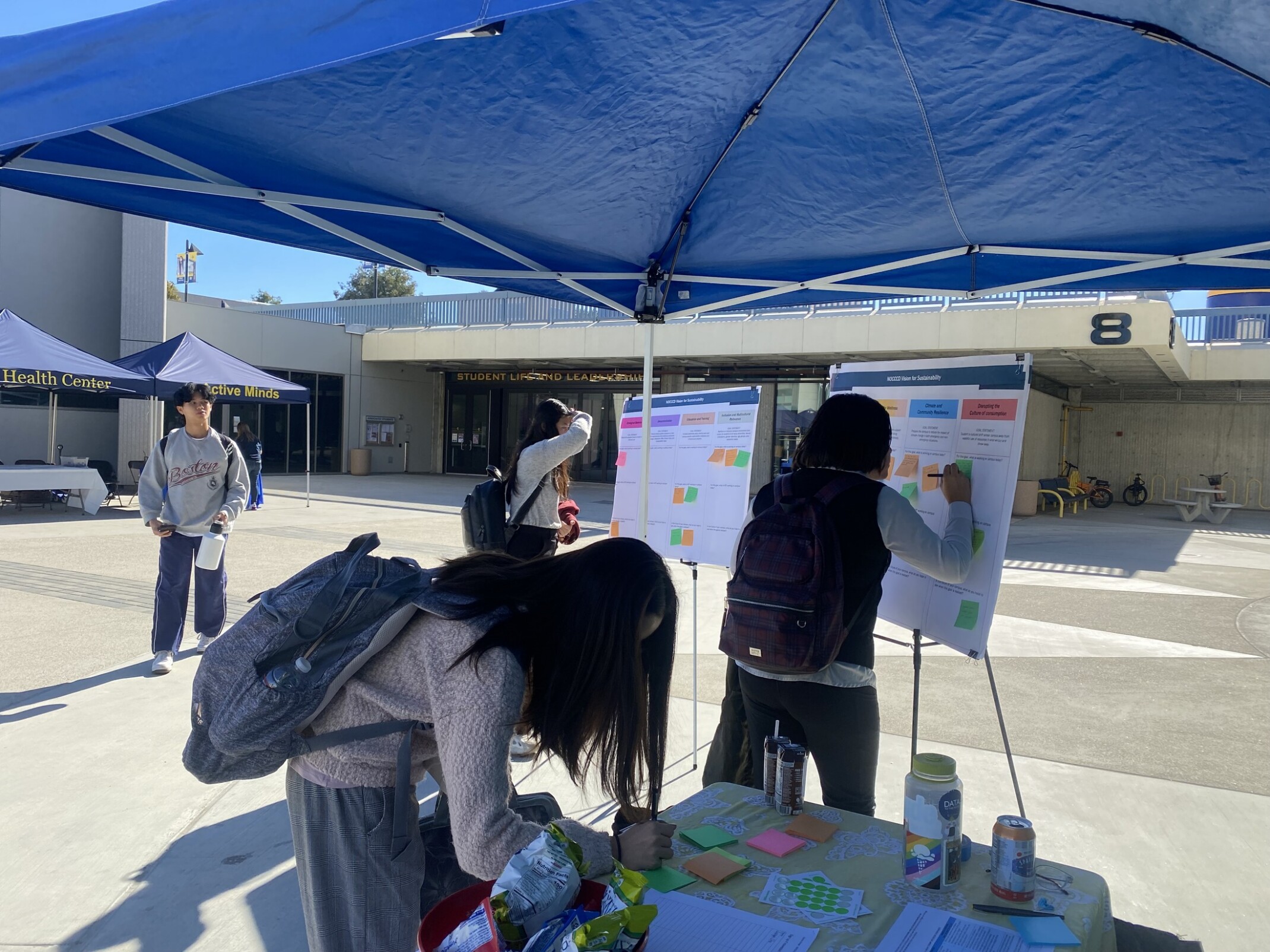 Students at an outdoor booth writing on sticky notes and placing them on foam display boards.