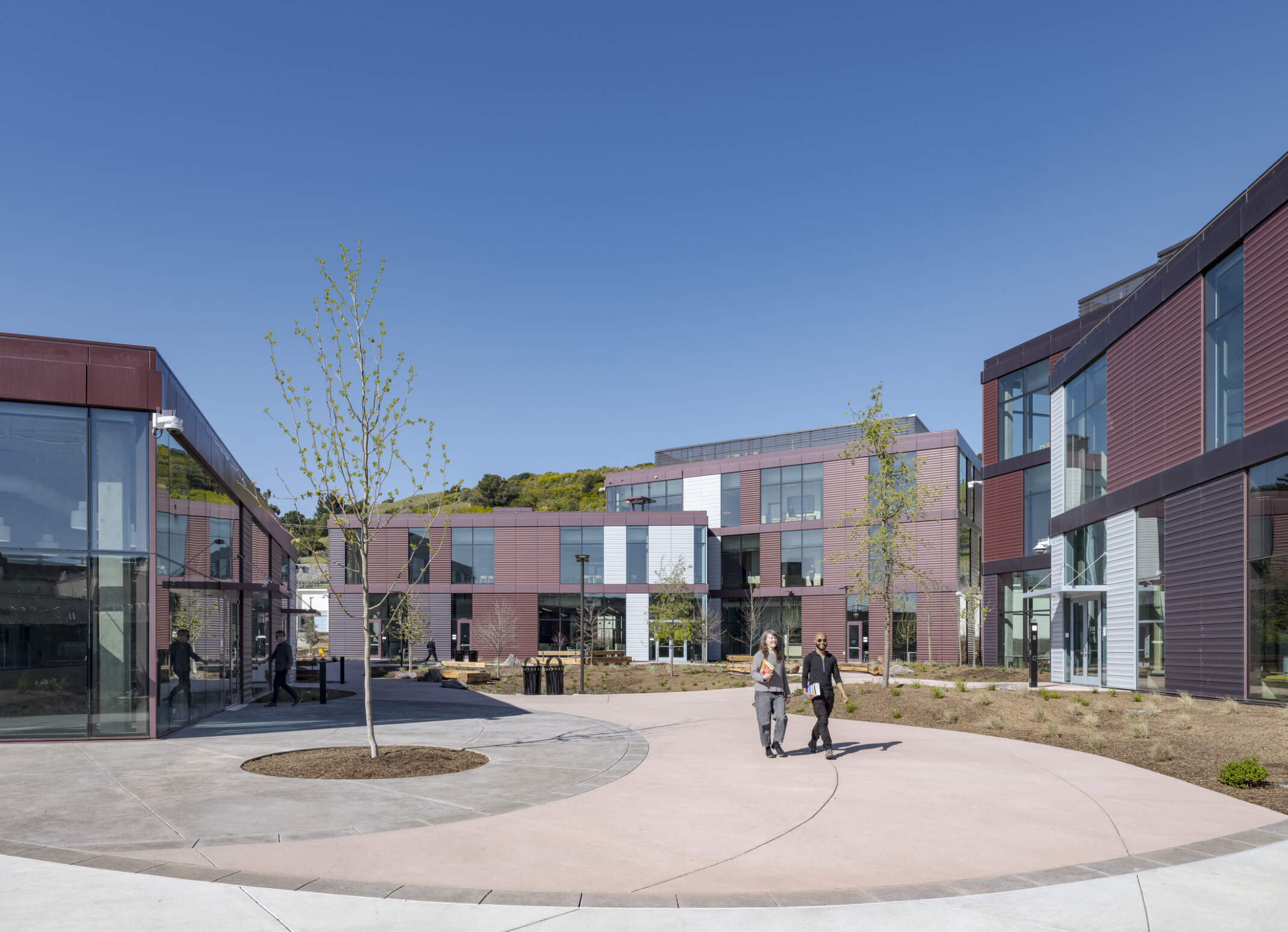 Wide shot showing the new center buildings in the background, with a courtyard in the foreground featuring wide walkways, natural landscaping, and people walking throughout.