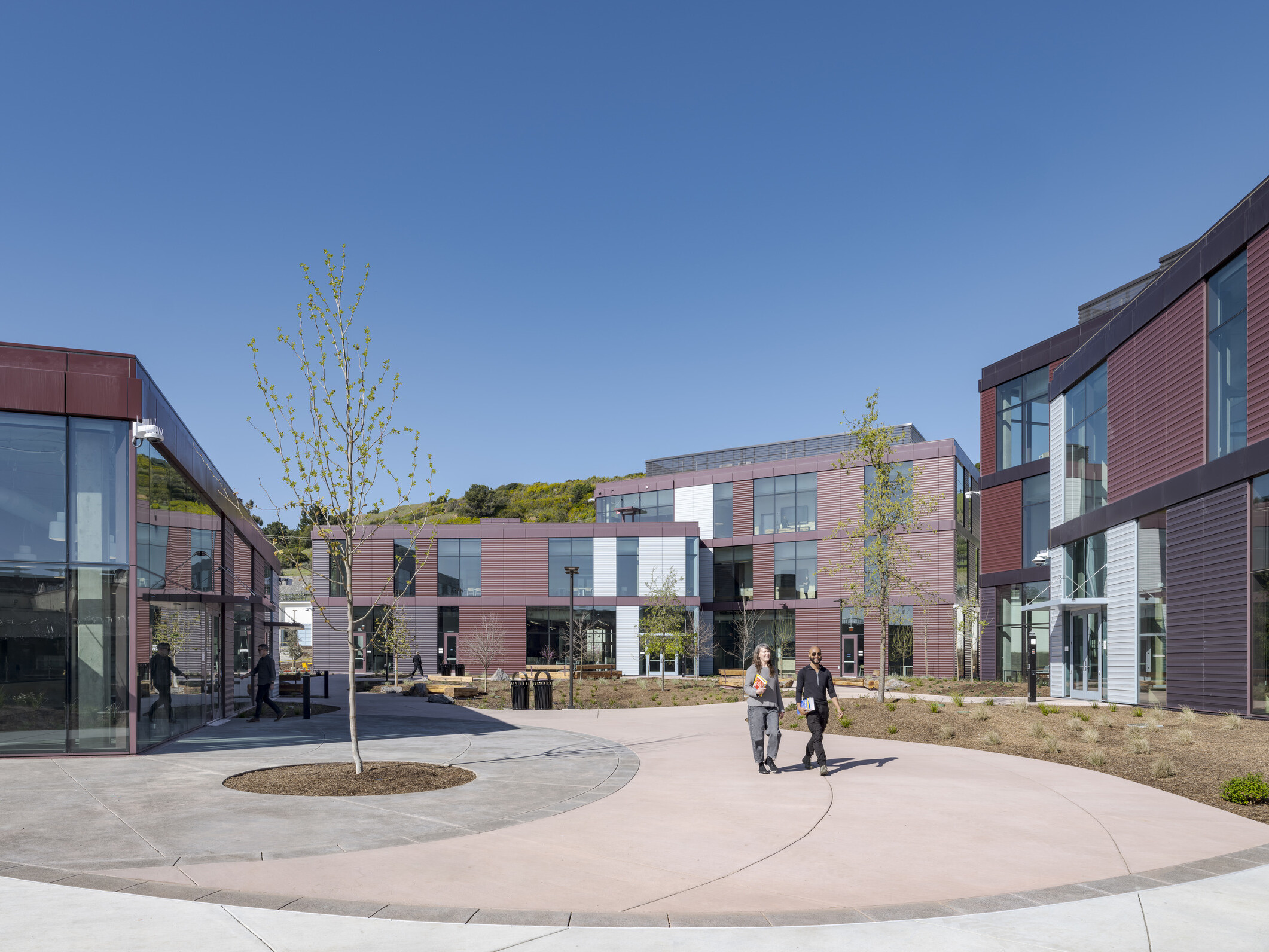 Wide shot showing the new center buildings in the background, with a courtyard in the foreground featuring wide walkways, natural landscaping, and people walking throughout.
