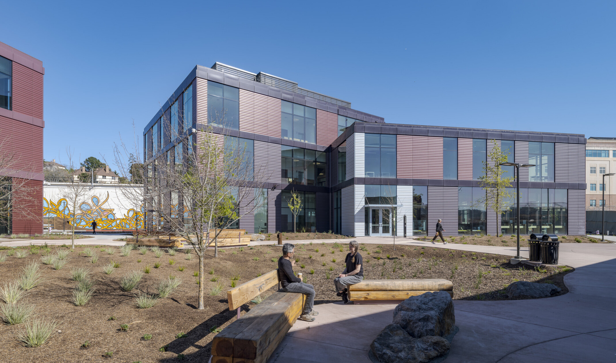 People sit chatting on natural wooden benches while one of the center's buildings stands in the background, with natural landscaping and ample walkways.